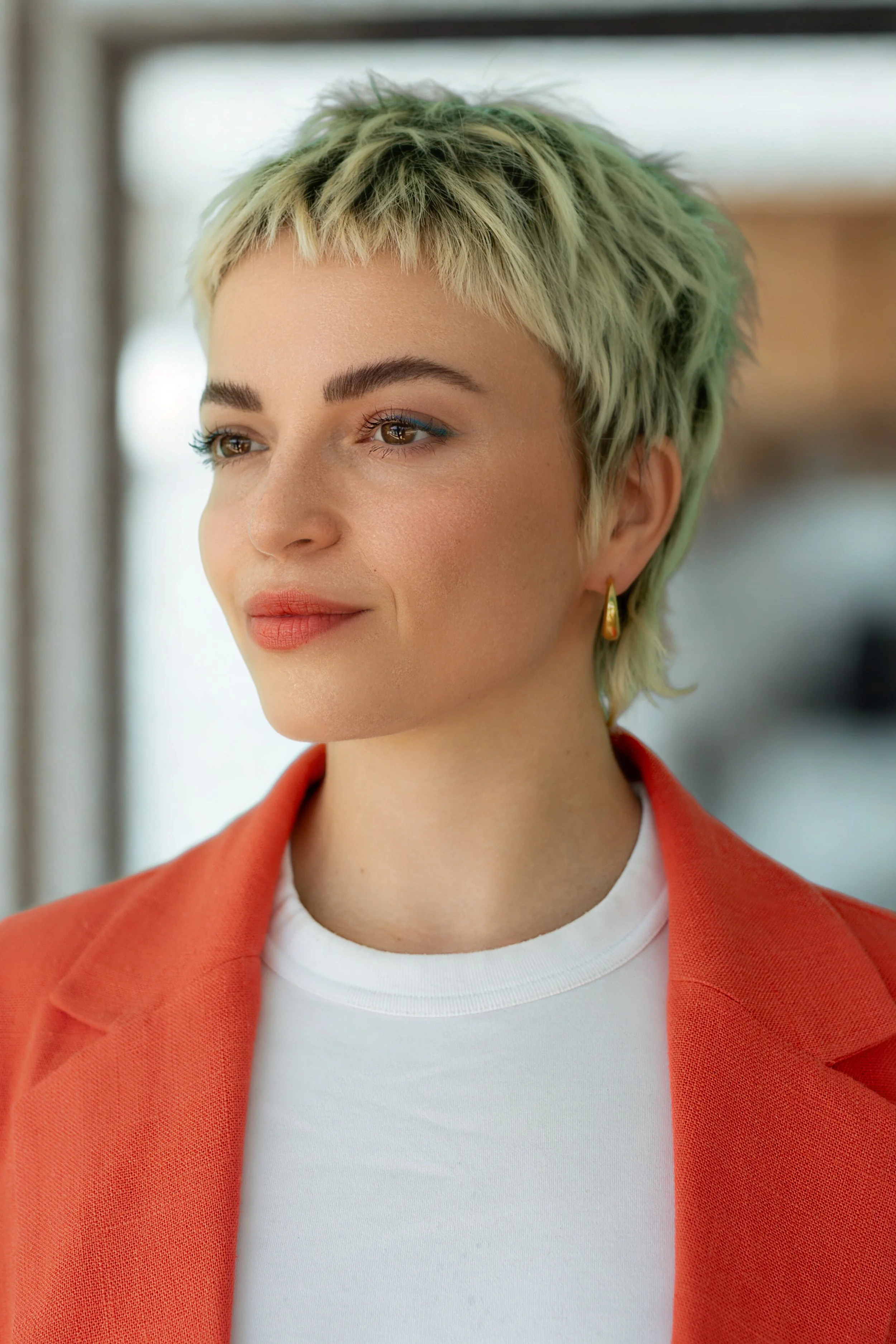 Close-up of a woman with short, blonde, textured hair wearing red blazer, white t-shirt, and gold hoop earrings, with a blurred window in the background.