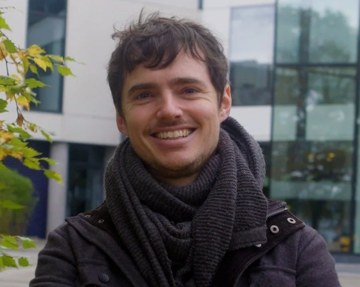 A smiling young man with brown hair and a scarf, standing outdoors in front of a modern glass building with trees.