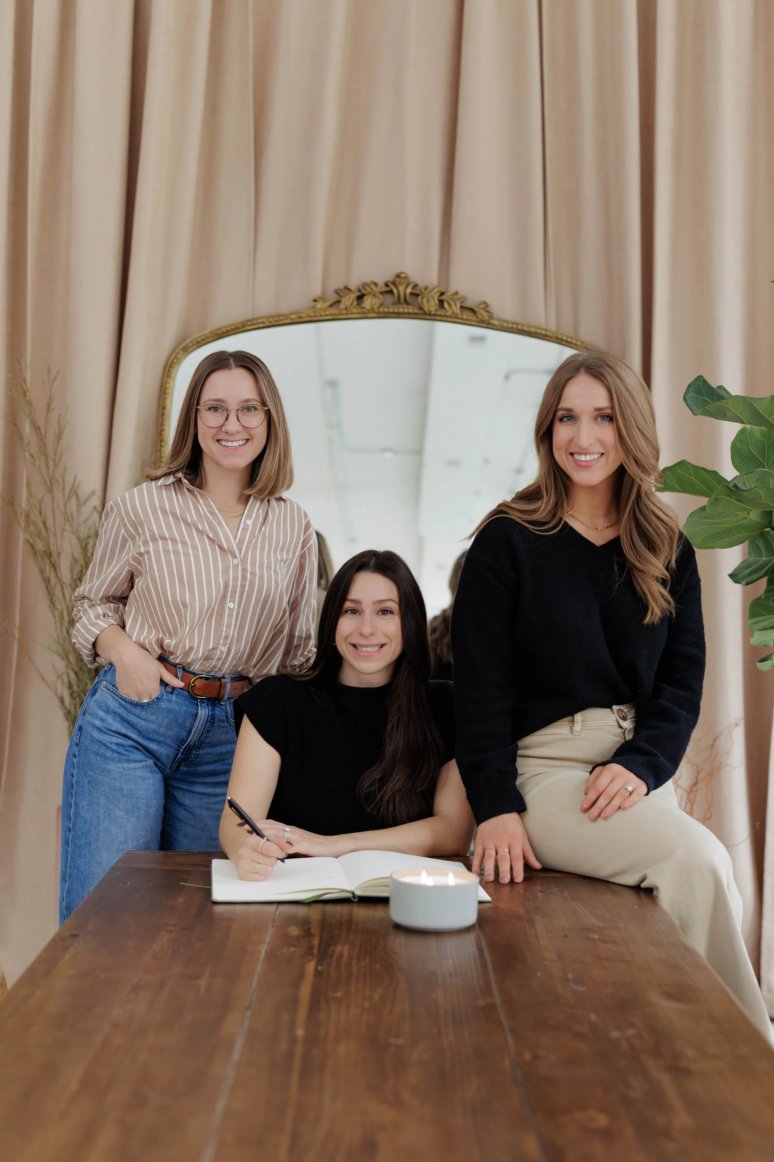 Three women in a stylish indoor setting with one writing in a notebook at a wooden table, a large mirror in the background, and a lit candle on the table.