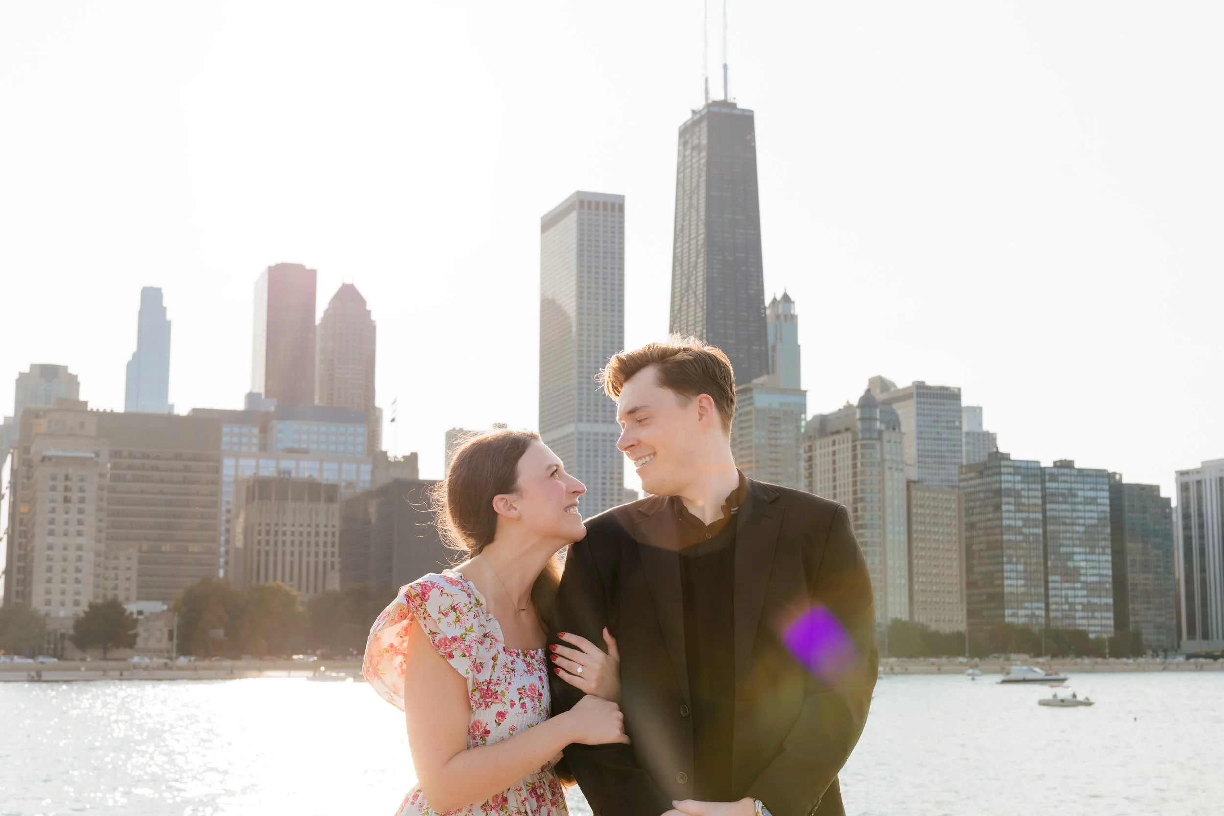 A smiling couple stands by the water with a city skyline, including a tall, distinctive building, in the background.