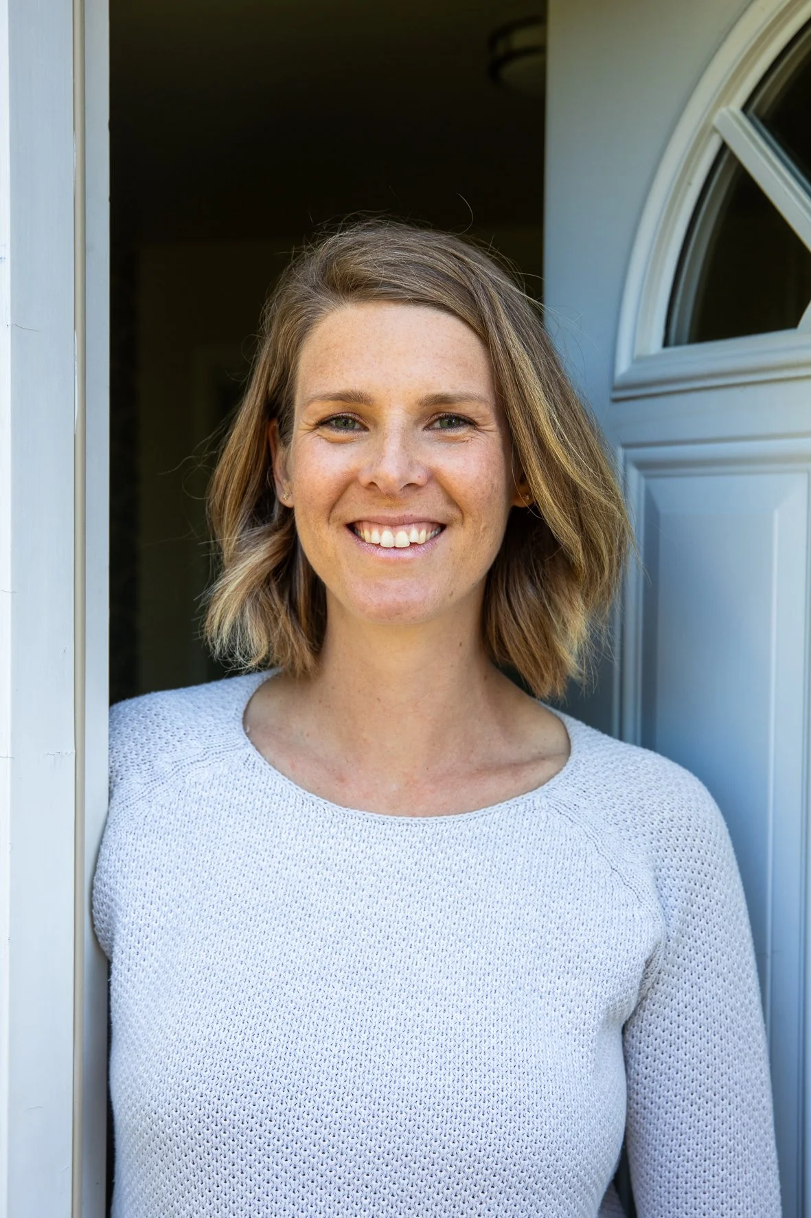 A woman with shoulder-length light brown hair and freckles smiling while standing in a doorway.