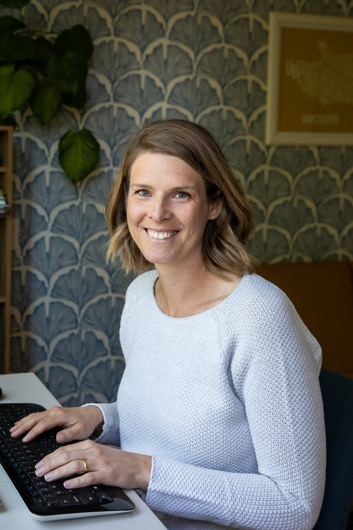 A woman sitting at a desk, smiling, typing on a black keyboard, with a patterned blue wallpaper background and green plants behind her.