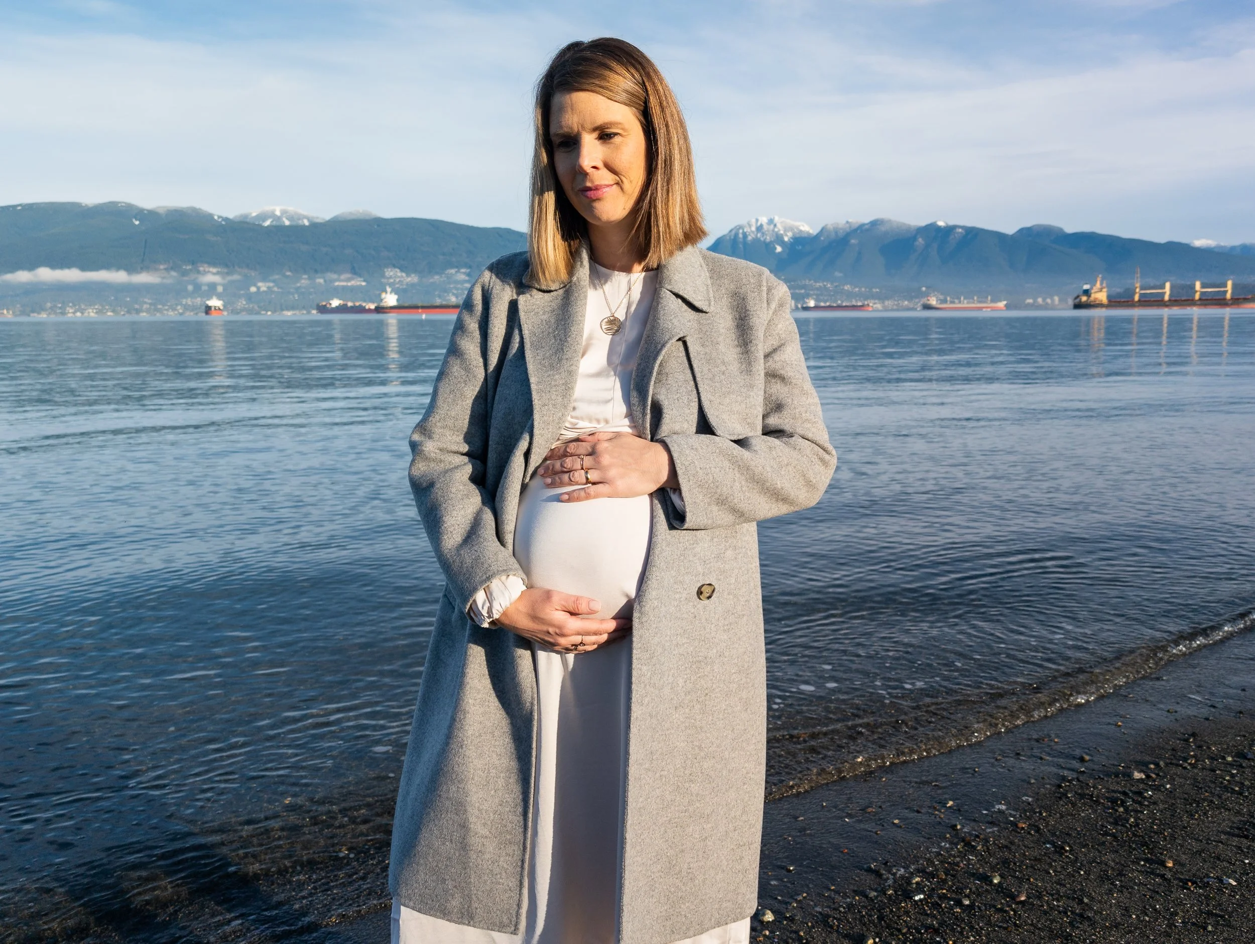 Verena on the beach with mountains in the background, holding her belly and looking contemplative.