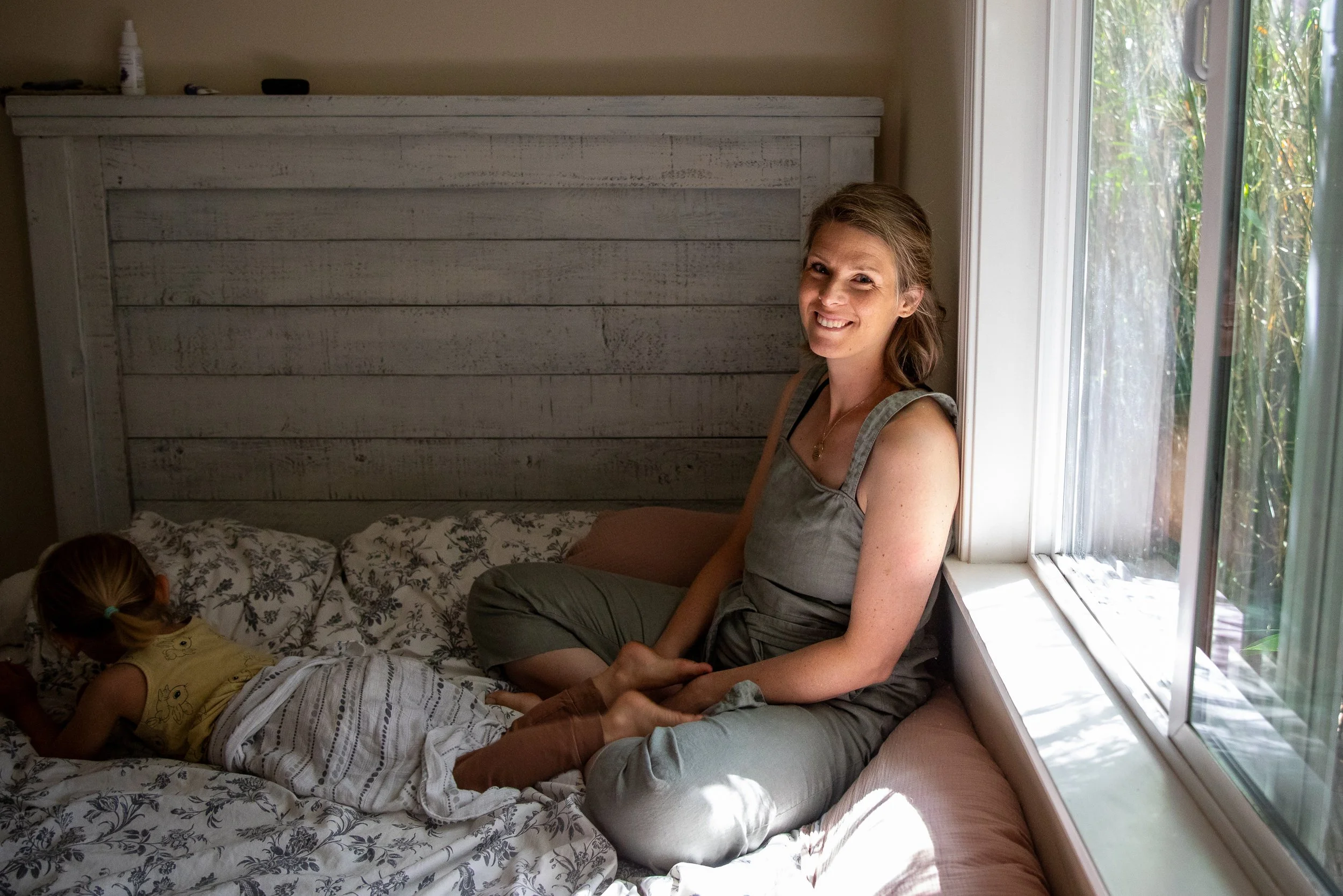 Verena sitting on a bed near a window, smiling, with a young child lying on the bed nearby, warm sunlight streams through the window.