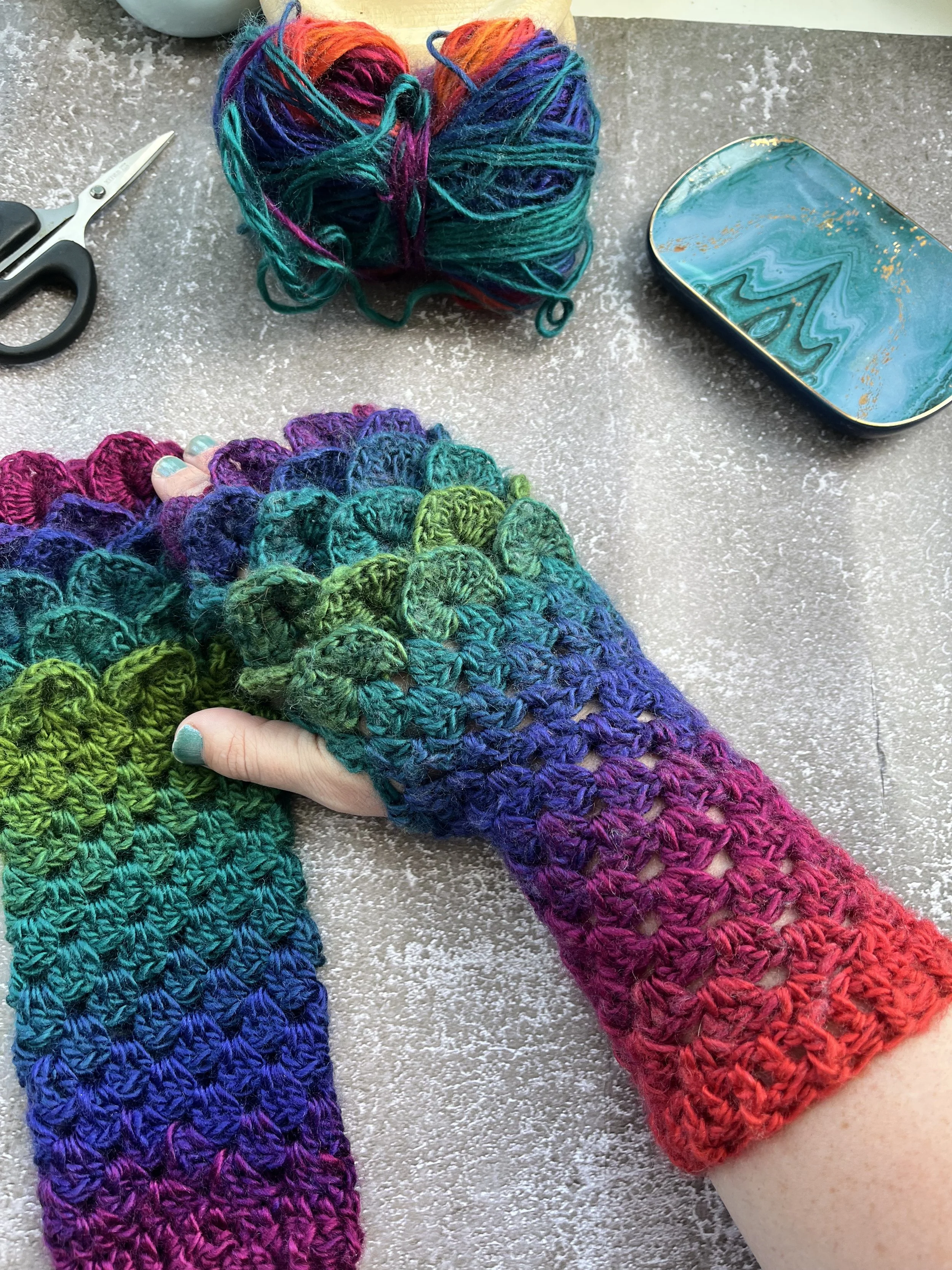 A uniques pair of handmade crochet dragon scale type fingerless gloves in unique blend of purple, blue, teal, green and red. Close up of dragon scale effect