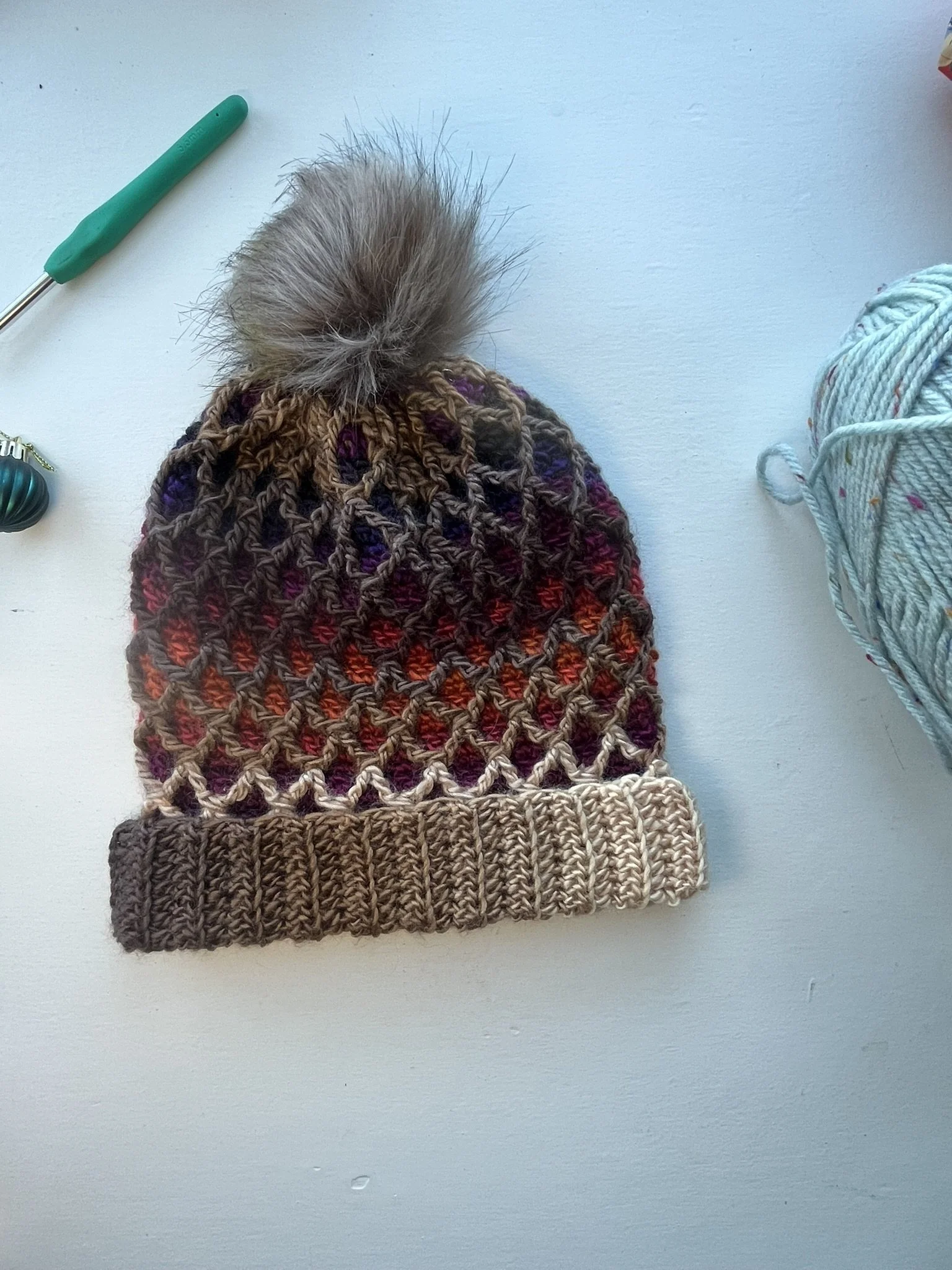 Overhead shot of a crochet beanie hat with a 3D diamond pattern in brown variegated yarn and a tan pompom