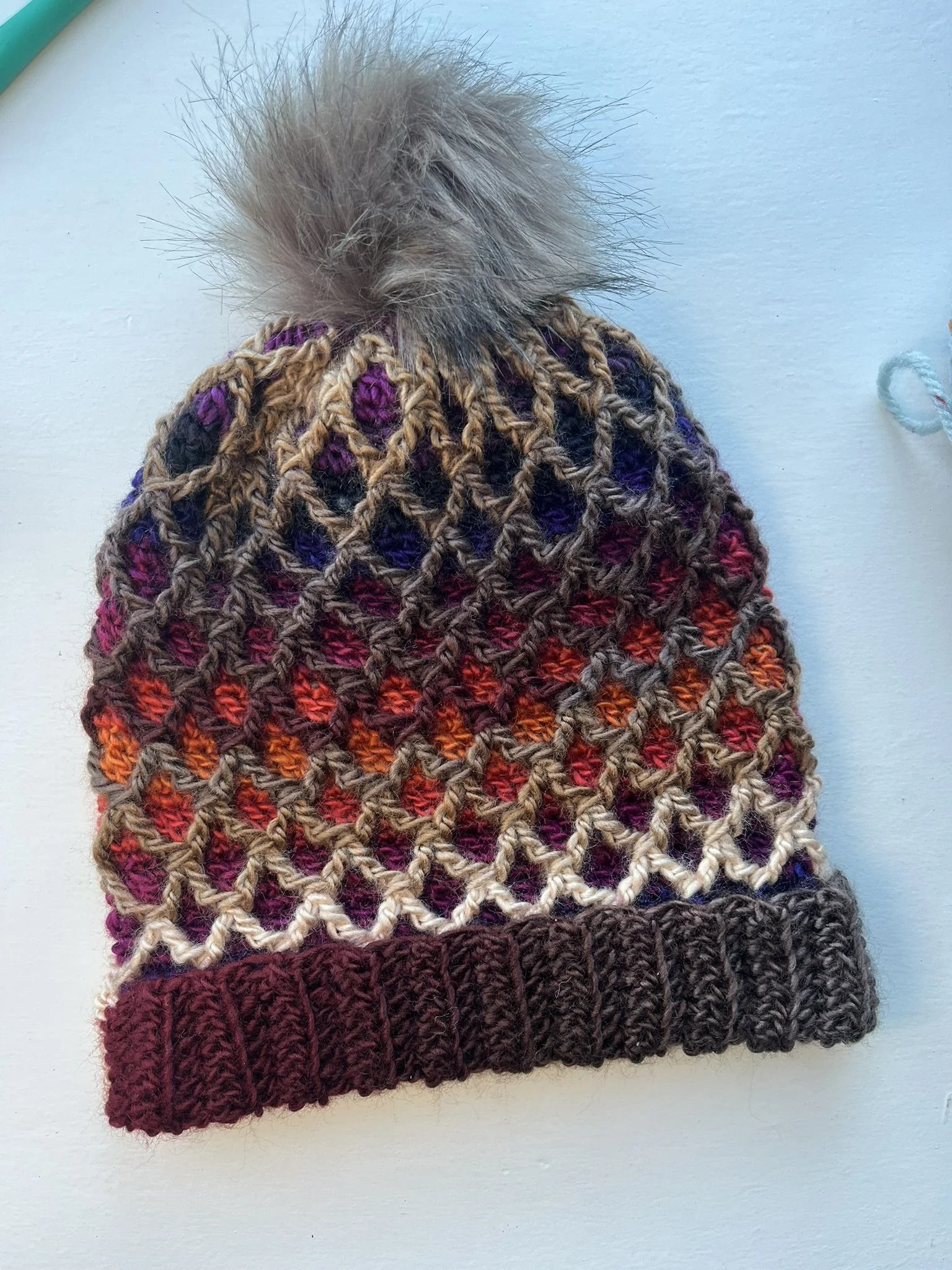 Overhead shot of a crochet beanie hat with a 3D diamond pattern in brown variegated yarn and a tan pompom