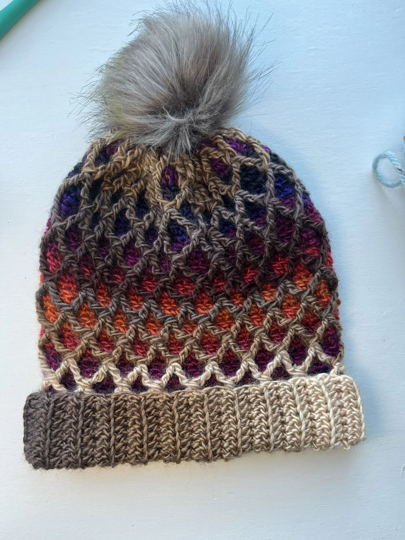 Overhead shot of a crochet beanie hat with a 3D diamond pattern in brown variegated yarn and a tan pompom