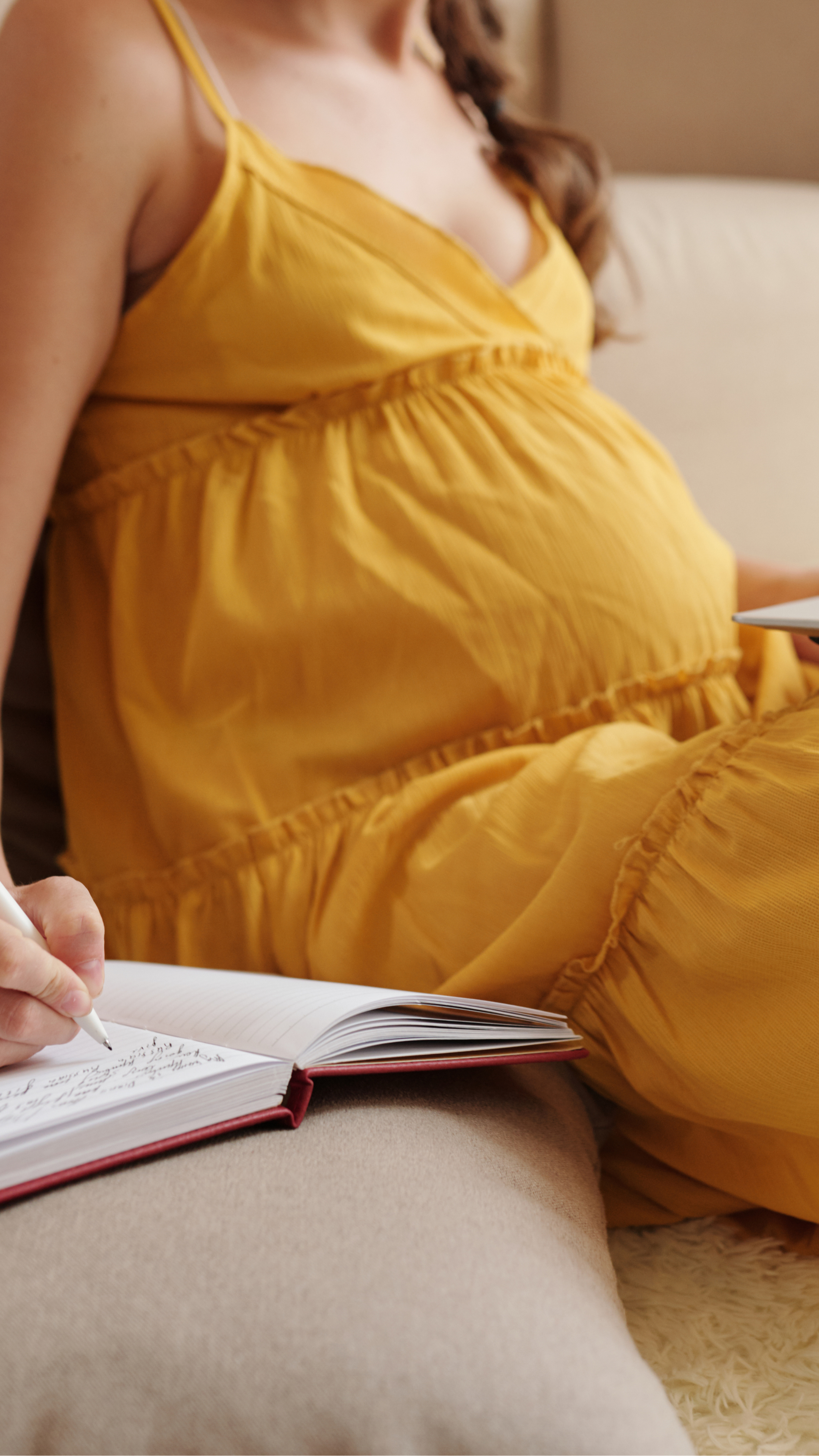 A pregnant woman in a yellow dress sitting on a beige couch writing in a notebook.