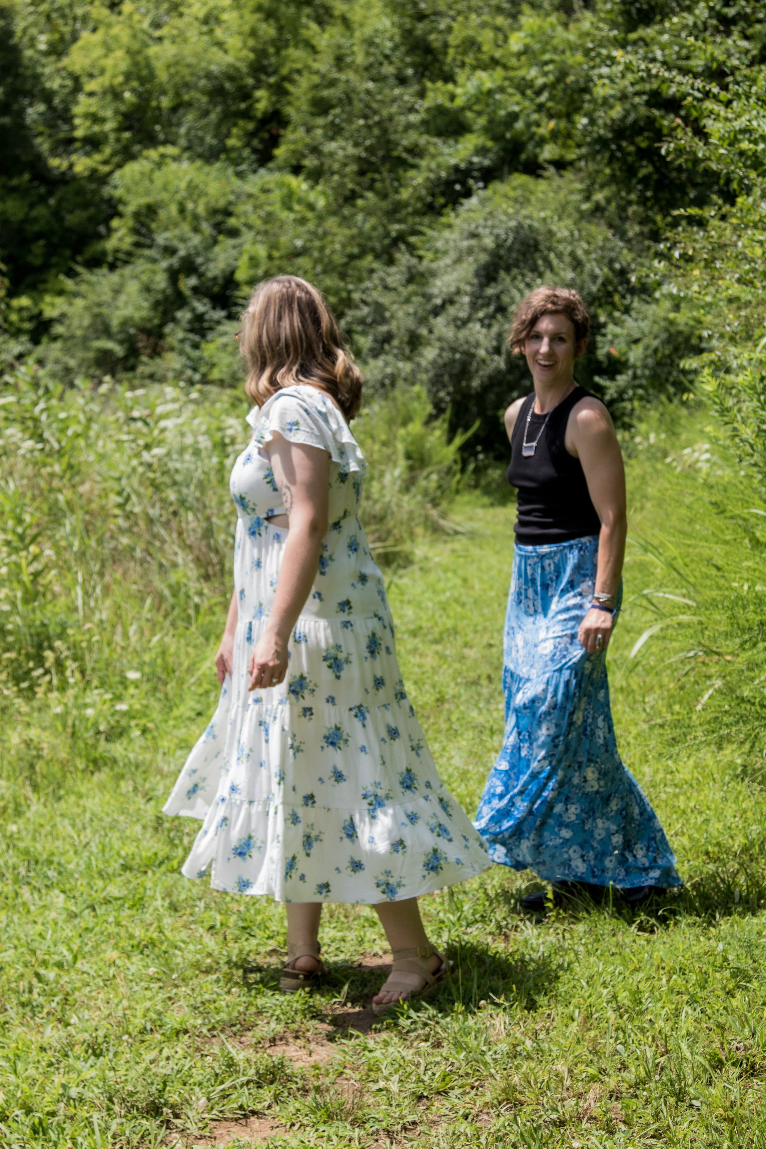 Two women, mothers and doulas walking outdoors on a grassy path surrounded by green trees and bushes, one smiling and looking at the camera, the other looking away.