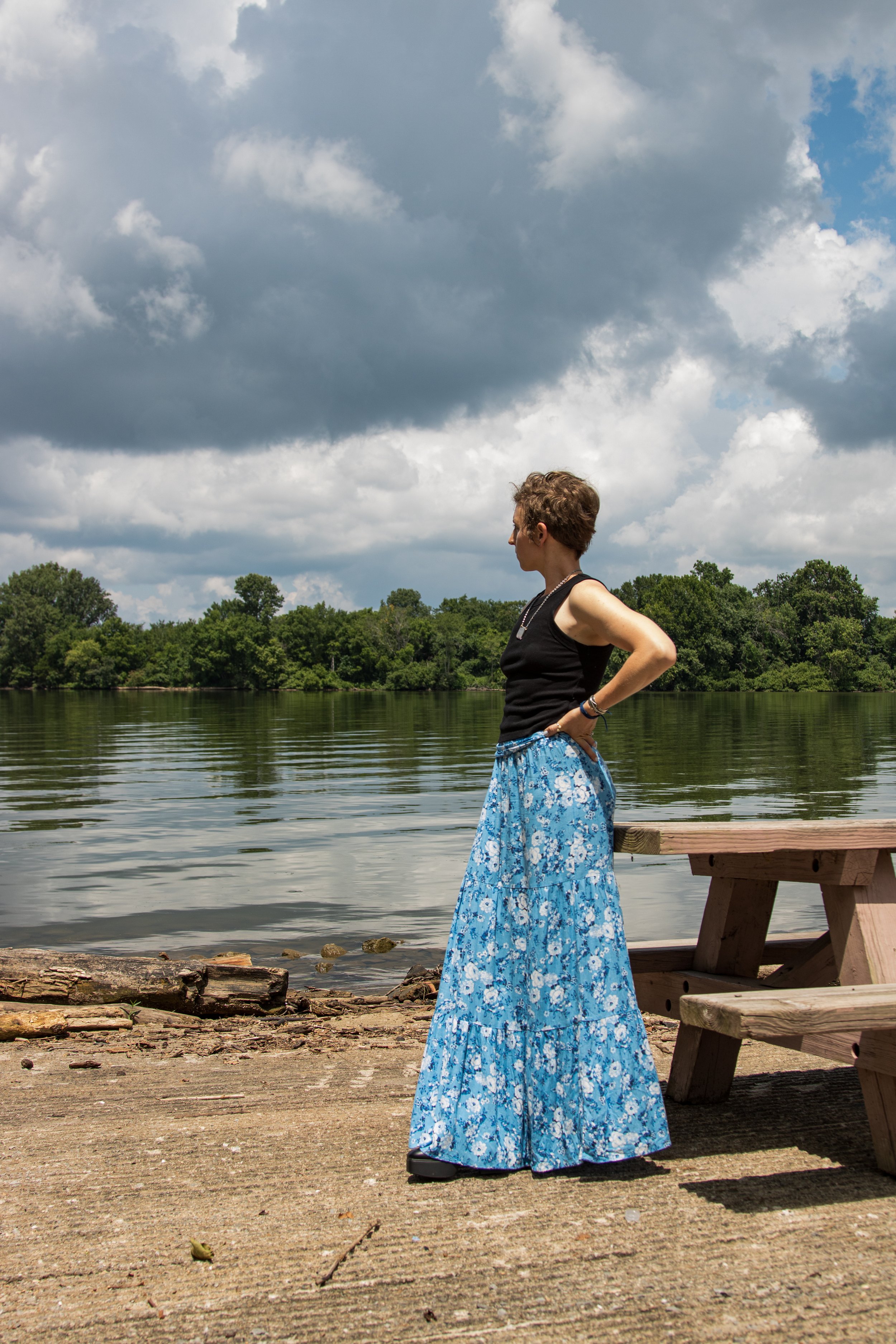Mallory Hansen owner of Mama Expanded. A woman standing on a concrete shoreline beside a wooden picnic table, looking out over a calm river with trees in the background, under partly cloudy skies.