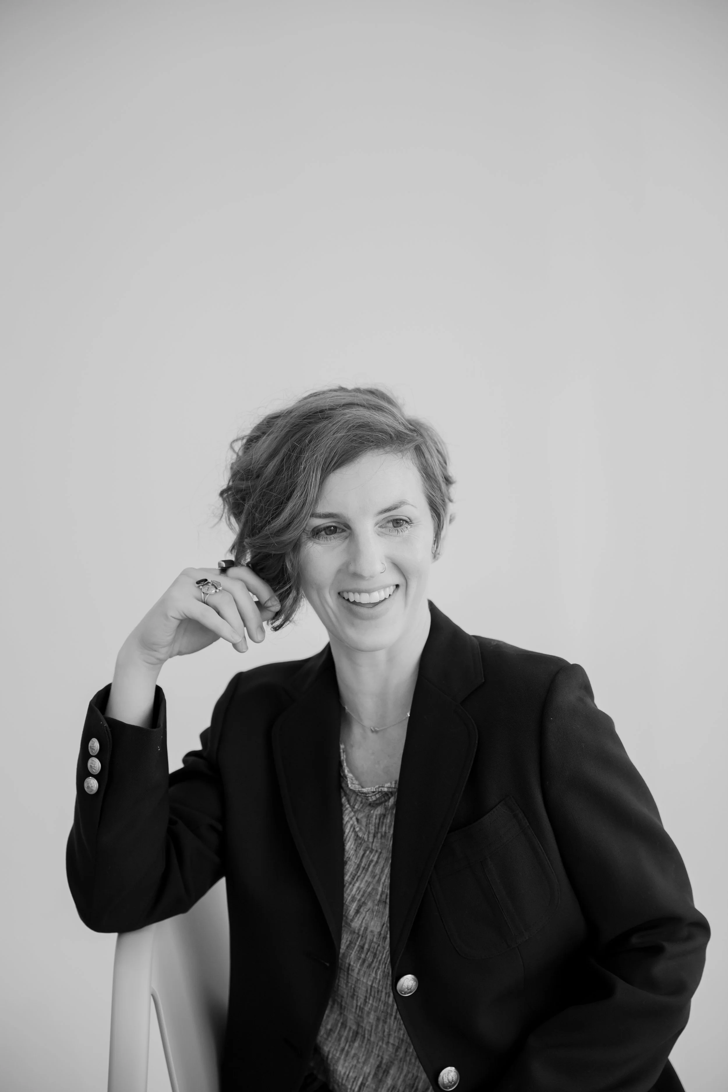 A woman with short, wavy hair smiling while sitting on a chair, wearing a dark blazer over a patterned shirt, against a plain background.