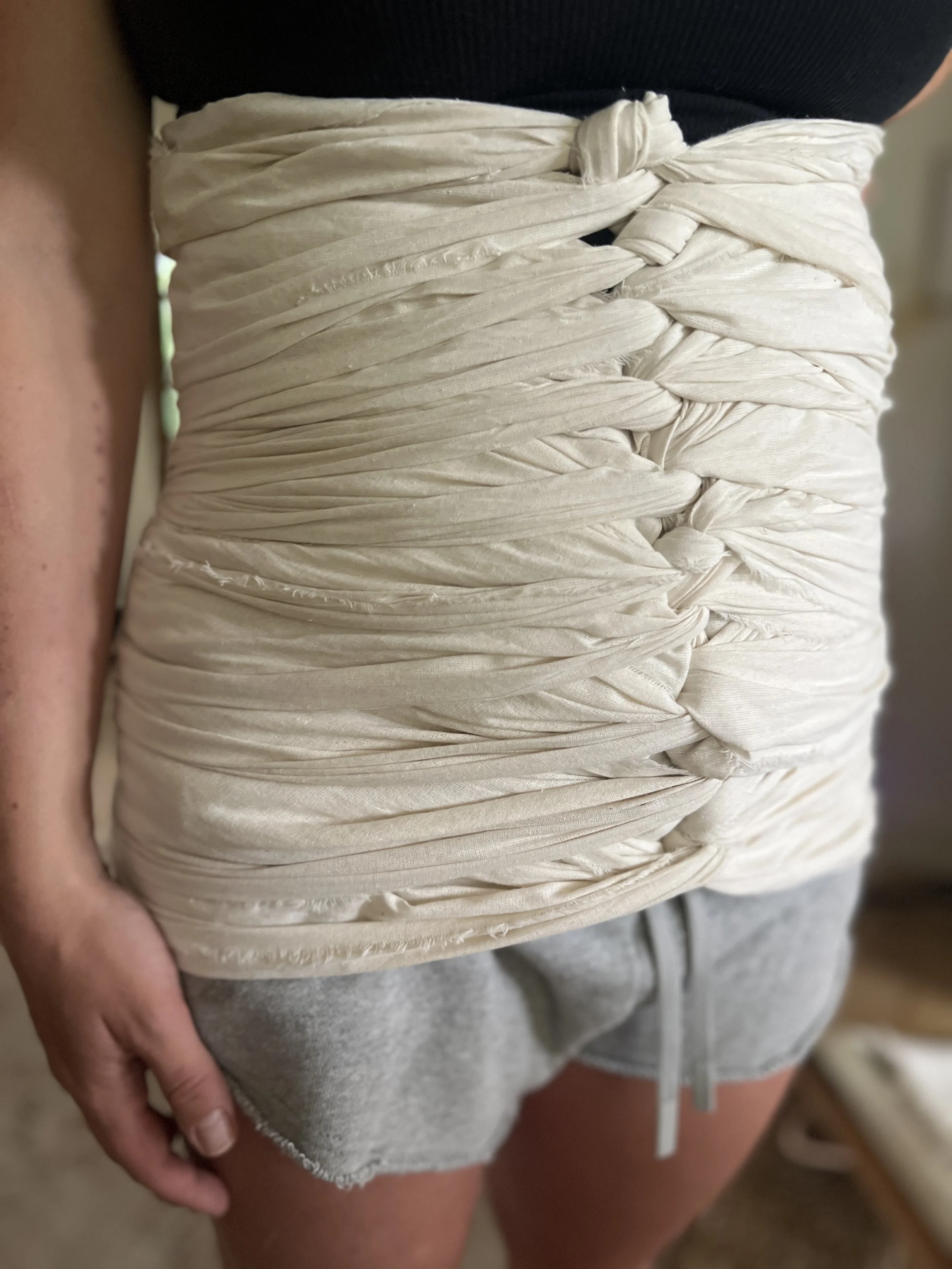 A person wearing a black top, with a cream-colored, ruched, and twisted fabric belt or wrap around their waist, paired with gray shorts.