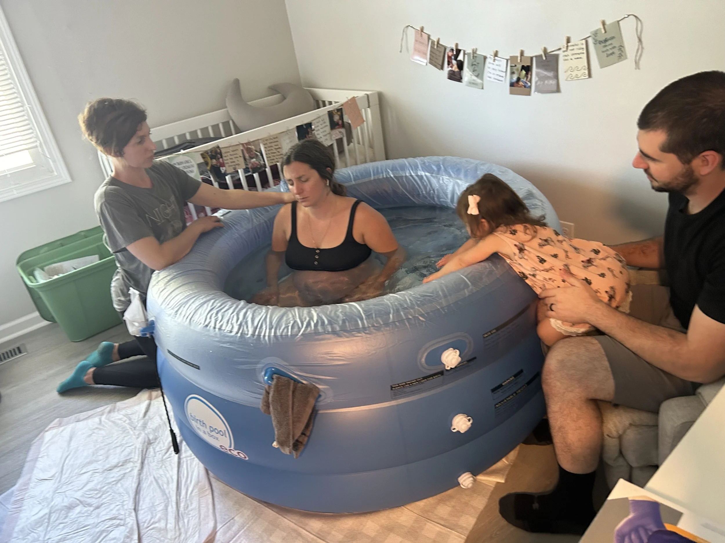 A woman is being baptized in a small inflatable pool with four people present. A man holds a young girl who reaches into the pool, and another woman supports the woman in the water. There are decorations and photos hanging on the wall behind them.