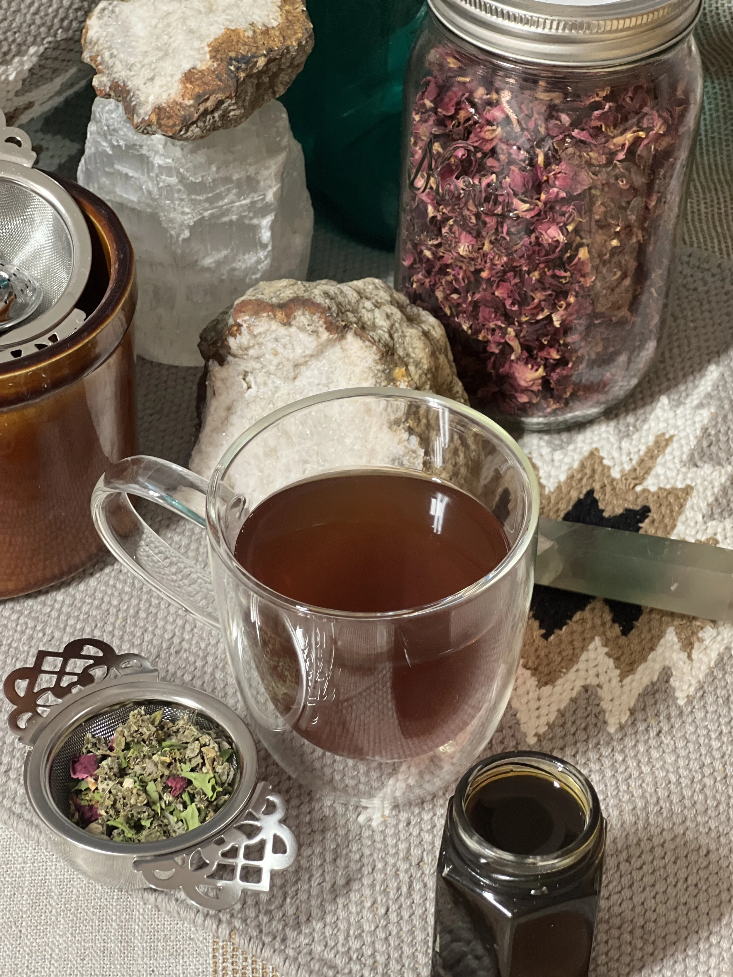 Mama Expanded Apothecary products, A glass mug filled with dark tea on a table surrounded by dried flower petals, rocks, a jar of dried flowers, crystals and various containers, with loose leaf tea and diffuser
