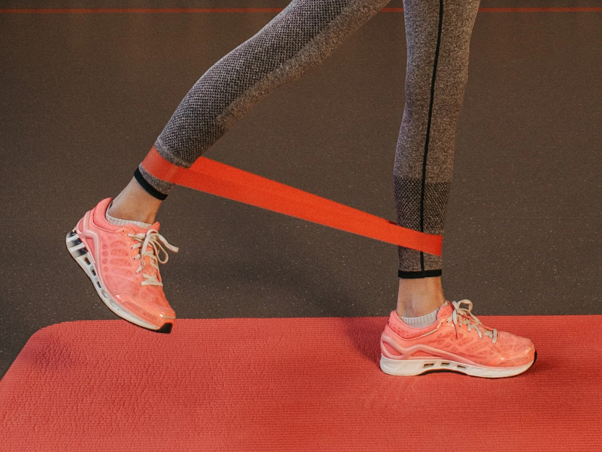 Person performing resistance band leg exercises on a red yoga mat, wearing pink athletic shoes, grey patterned leggings, and using an orange resistance band. Restore and Strengthen 6 week series for postpartum training.
