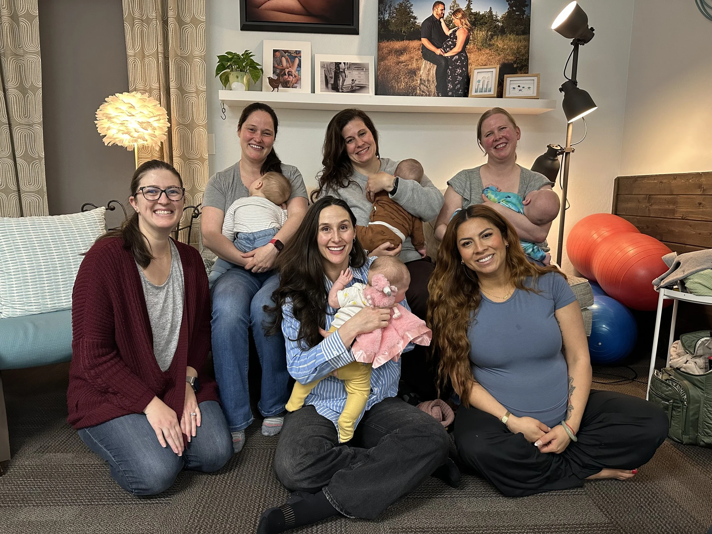 Two women each holding a baby, one woman sitting on a bean bag chair, and one woman standing and speaking with hand gestures in a room with chairs, a television, and wooden wall paneling.