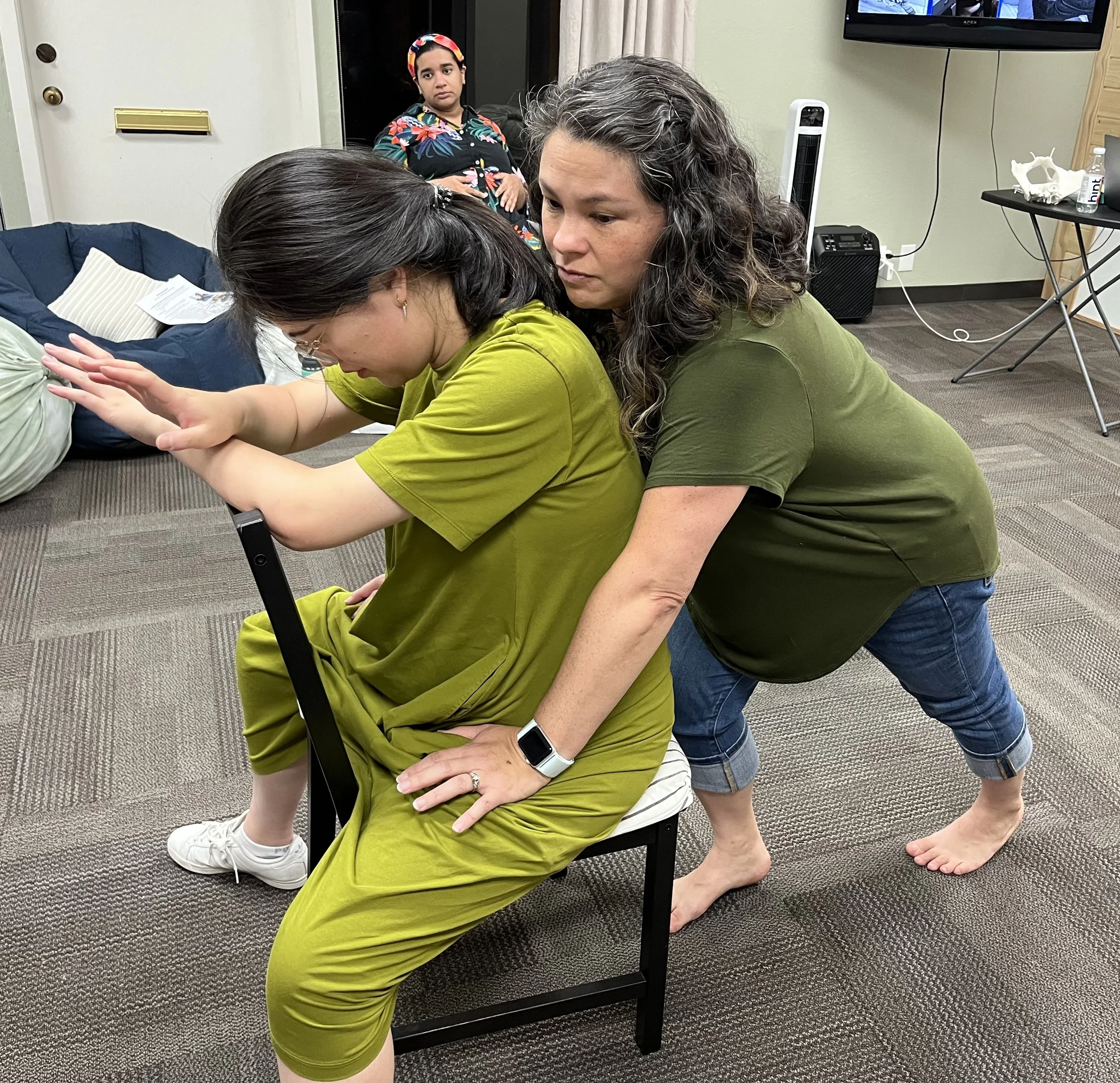 A woman with long brown hair, wearing a black shirt with a floral pattern, is smiling and leaning close to a seated woman with long dark hair, wearing a black tank top, who has her arms crossed. They are in a room with a wooden wall and a television screen showing images in the background.