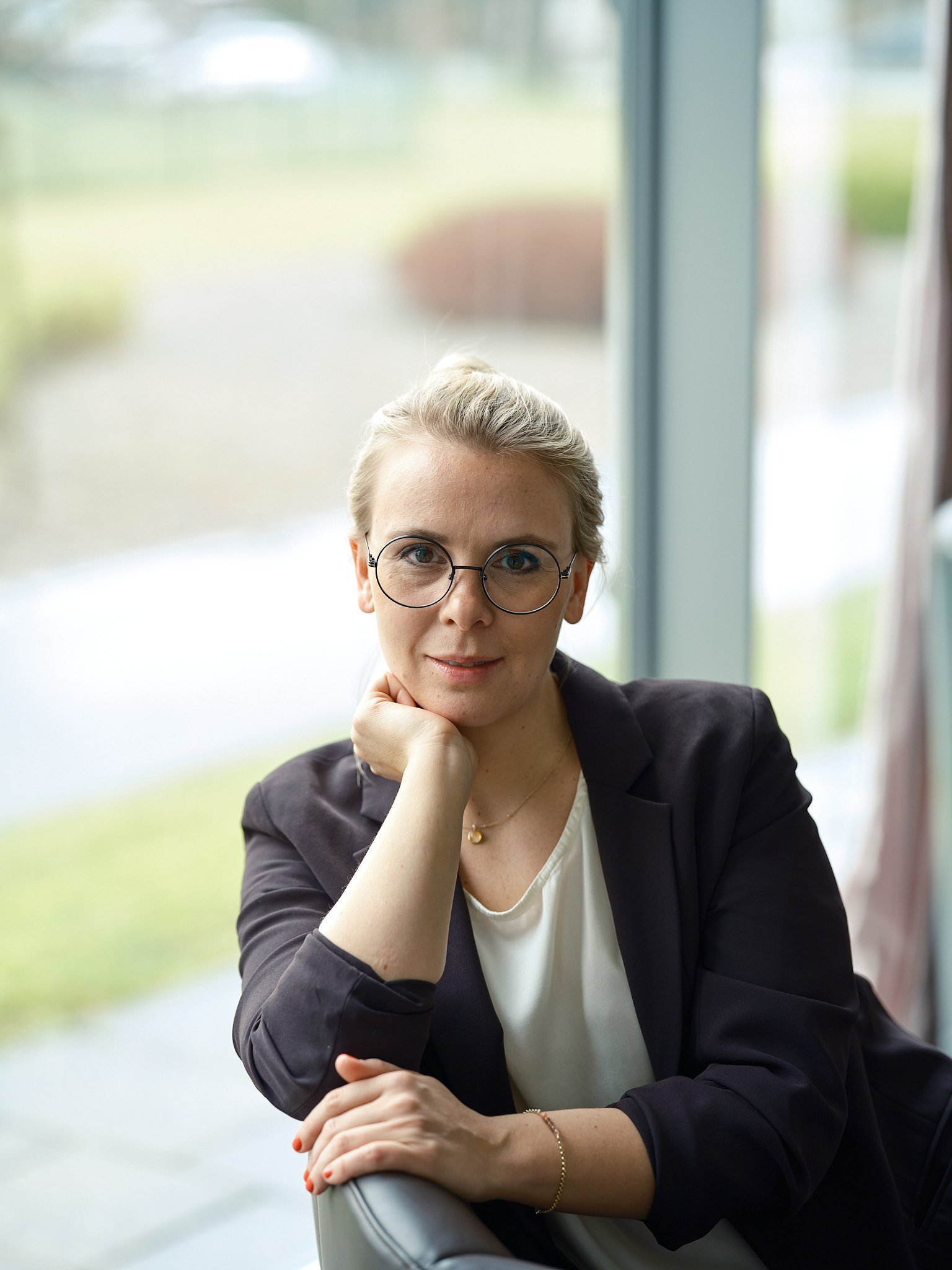 Nahaufnahme einer Frau mit kurzen blonden Haaren, Brille und schwarzem Blazer, die im Freien vor einem Fenster sitzt, lächelnd in die Kamera schaut.