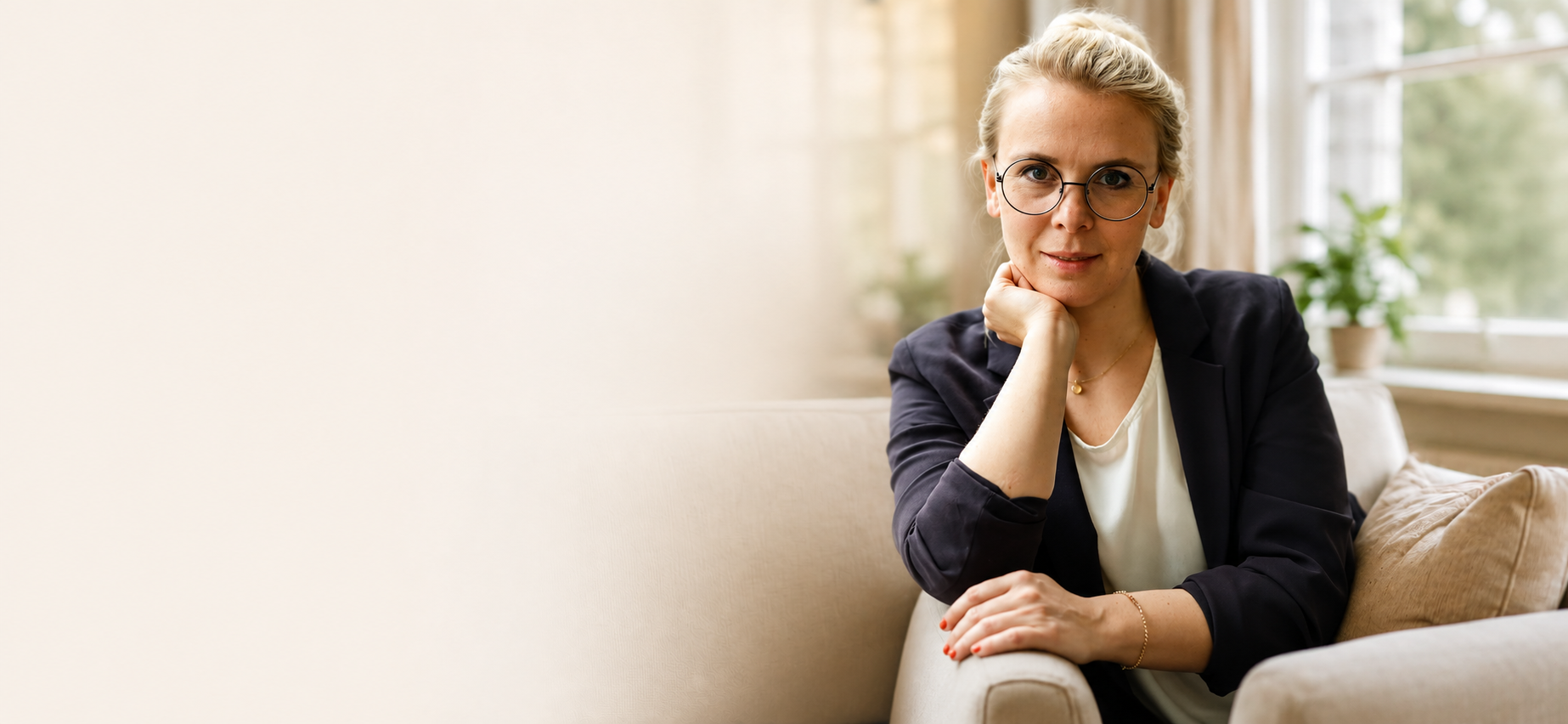 Frau mit Brille, dunkler Blazer, sitzend auf einem Sofa in einem hellen Raum, Blickrichtung zur Kamera, Hand stützt das Kinn, natürliche Beleuchtung, pflanzen im Hintergrund.