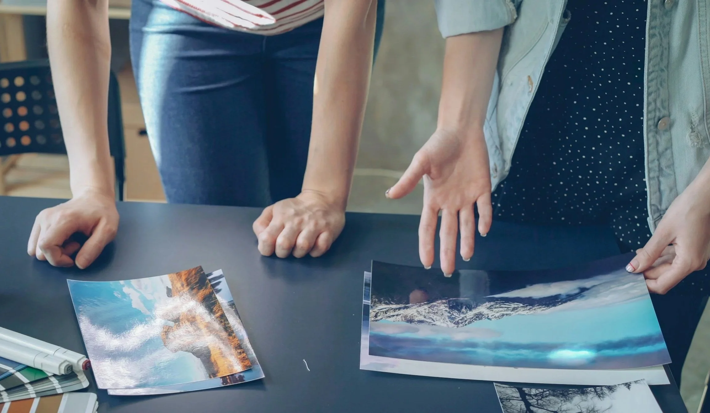 Two people are looking at large photographs or landscape images spread on a black table, with their hands pointing and touching the photos, in an indoor setting.