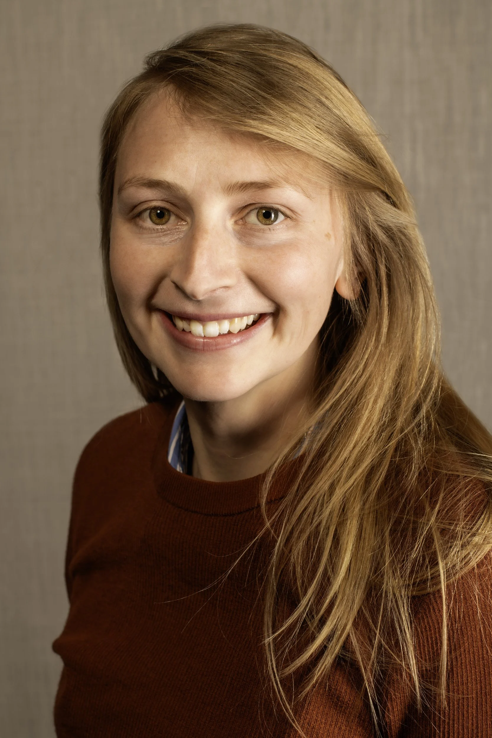 A close-up portrait of a woman with long blond hair, wearing a red sweater, smiling at the camera against a neutral background.
