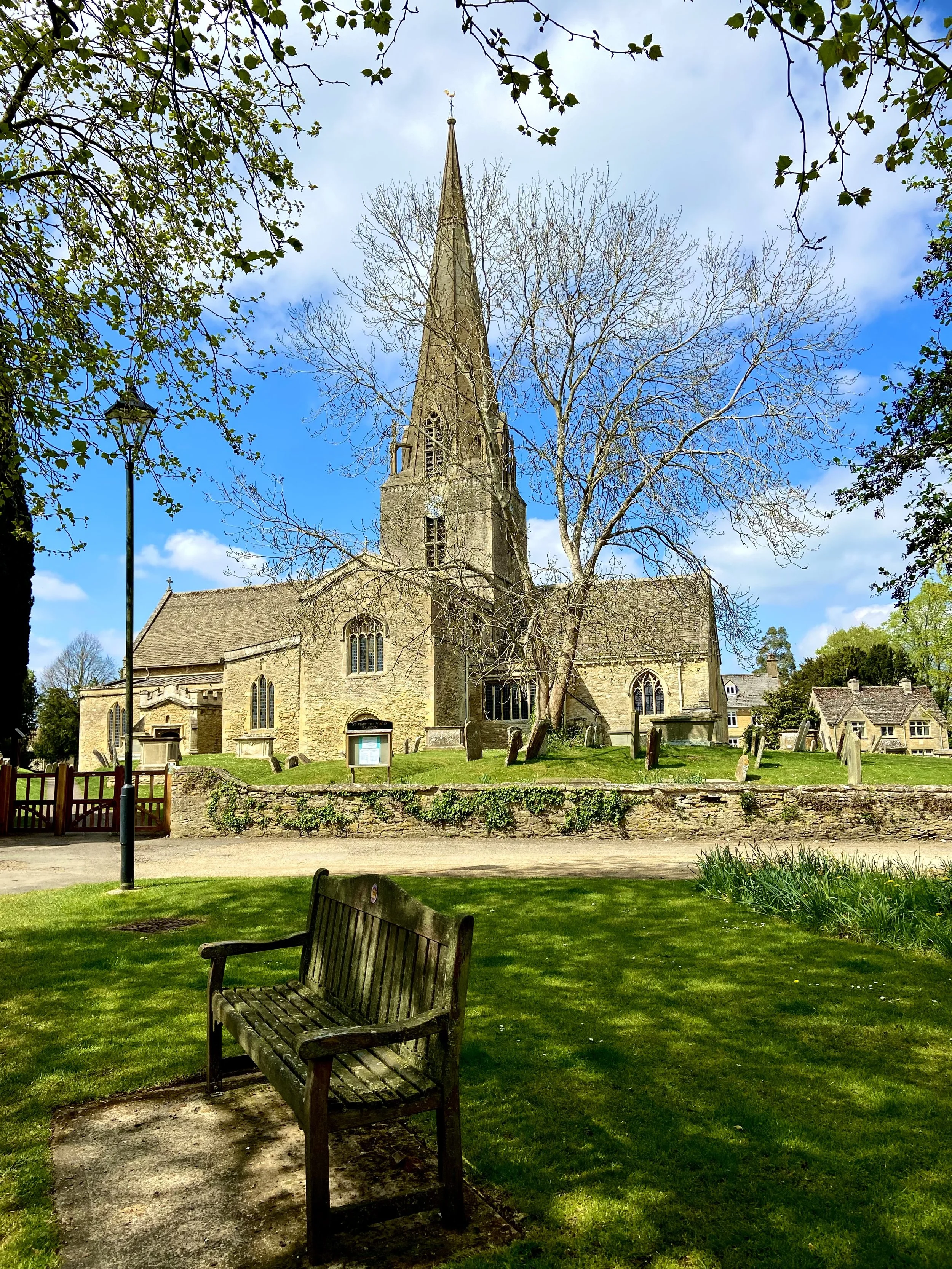 A historic, globally famous Cotswold stone church. Downtown Abbey was filmed in this 10th or 11th Century Church. It's tall steeple, represents the Gothic style architecture. Surrounded by a grassy area with trees, benches, and a low stone wall.