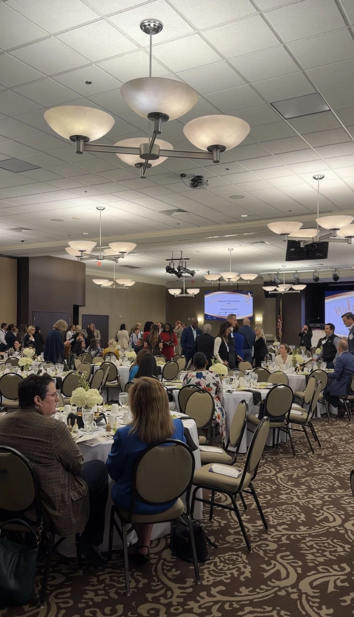 A large banquet hall filled with round tables set with white tablecloths, floral centerpieces, glassware, and place settings. People are dressed in formal attire, mingling, and sitting at the tables. A stage with a large screen displays a welcome mes