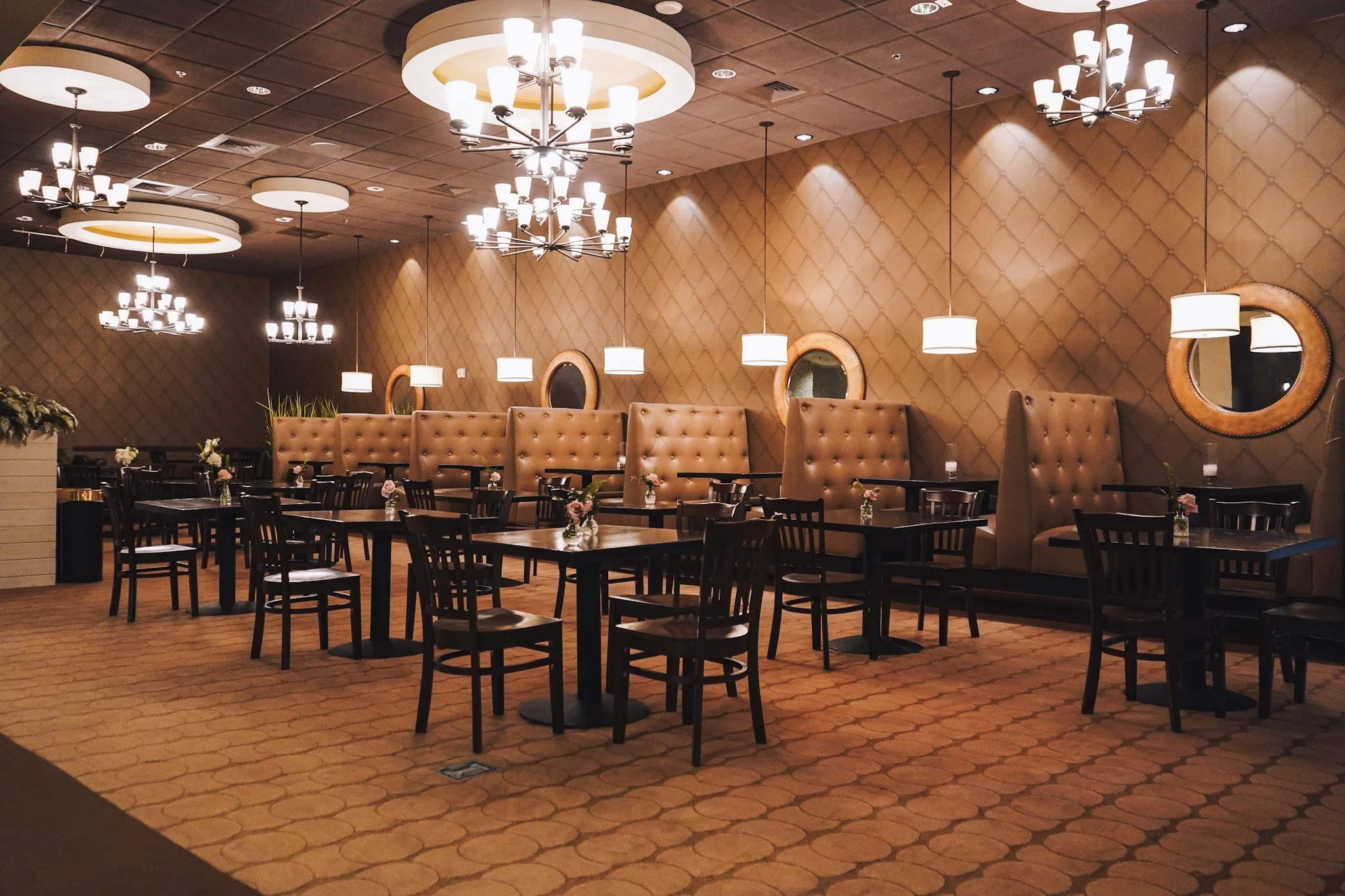 Elegant restaurant with high booths, wooden tables, black chairs, ambient lighting fixtures including chandeliers and pendant lights, and decorative mirrors on the beige quilted wall.