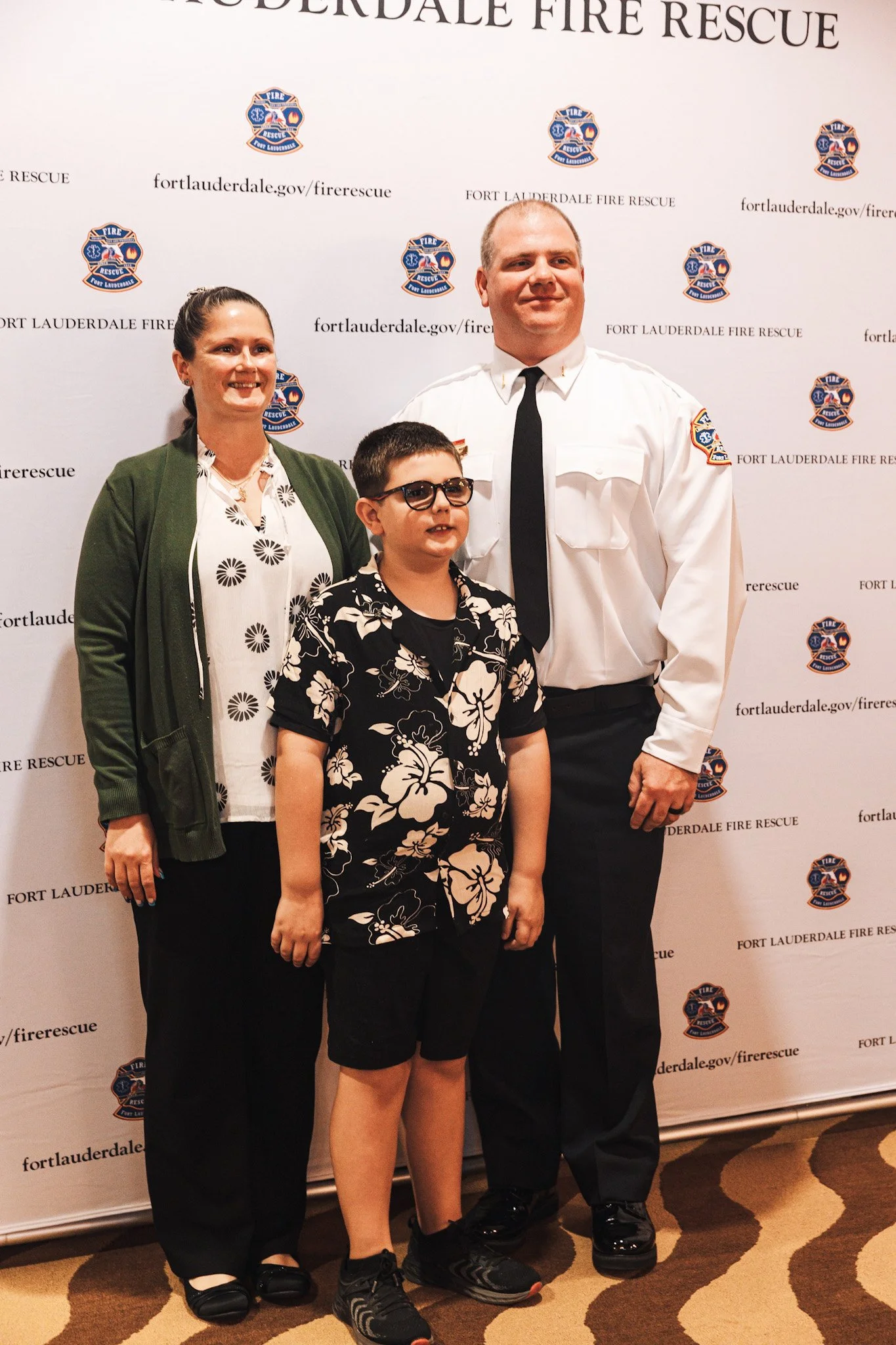 A group of three people, including a firefighter in uniform, a woman, and a young boy, standing in front of a backdrop with Fort Lauderdale Fire Rescue logos and website, smiling for a photo.