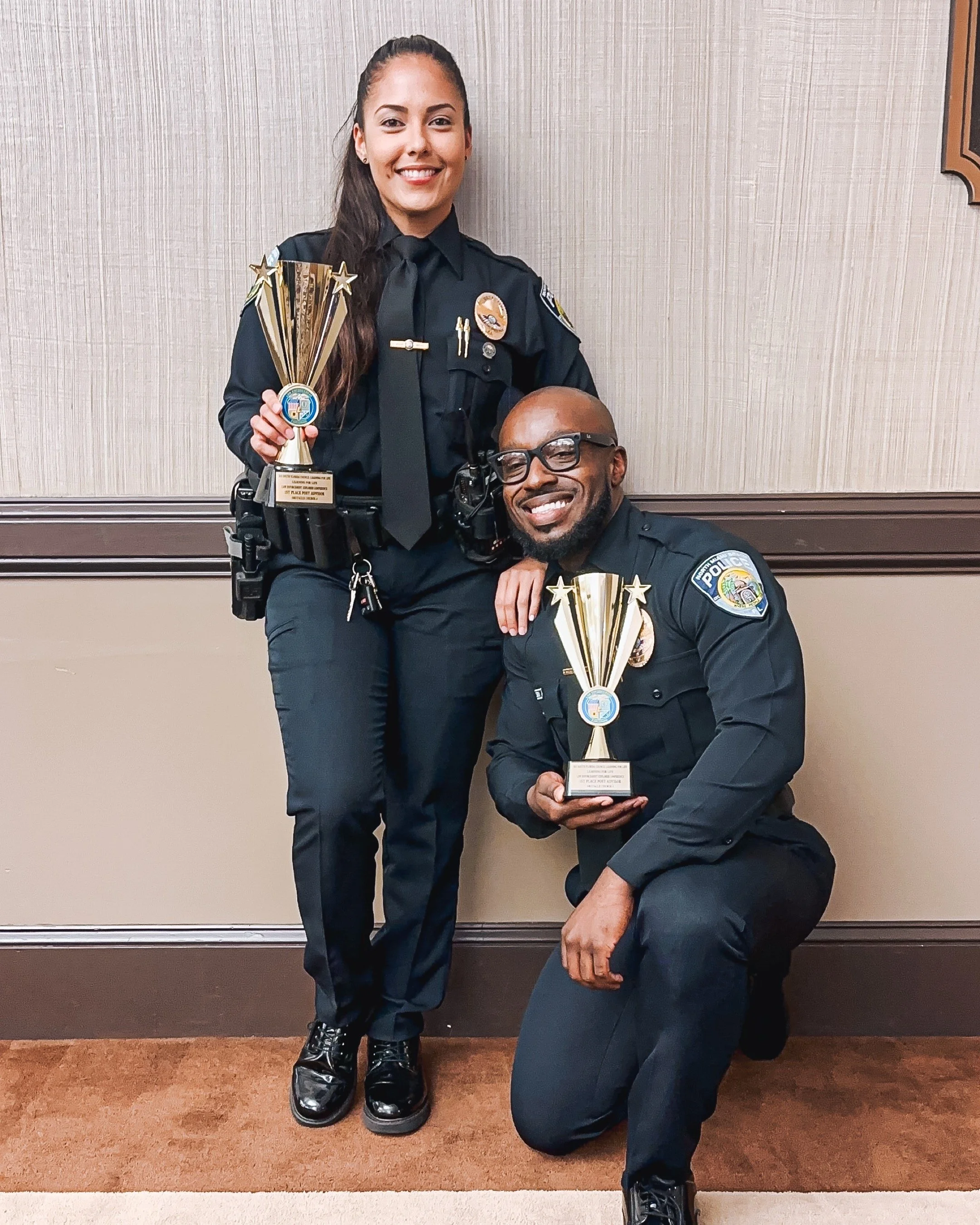 Two police officers, a woman and a man, smiling and holding trophies, in uniform, against a beige and brown wall.