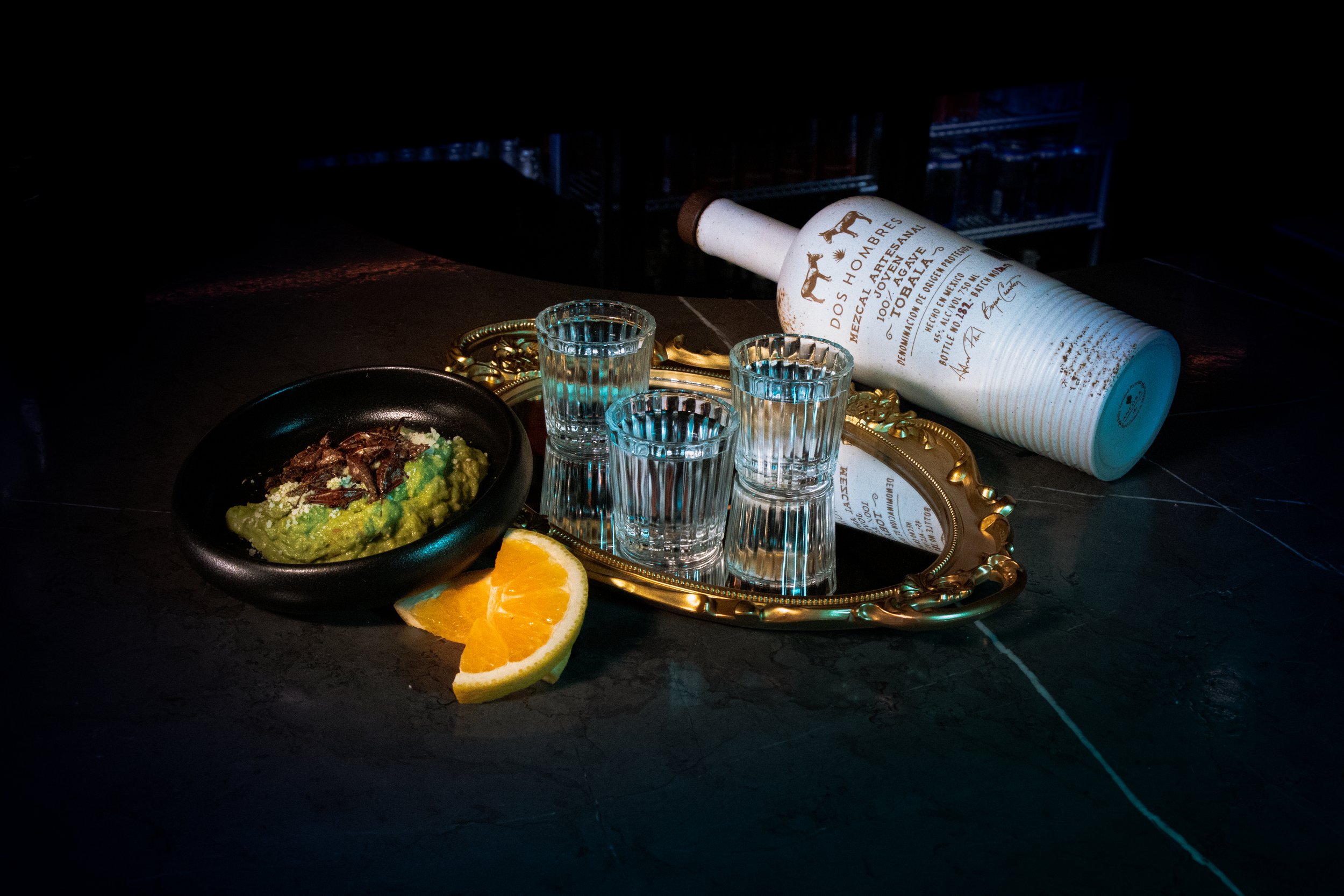 Tequila bottle with shot glasses on golden tray, guacamole in bowl, and orange slices on dark table. sabina mezcaleria boston, food photography, professional photographer boston, Mexican restaurant boston