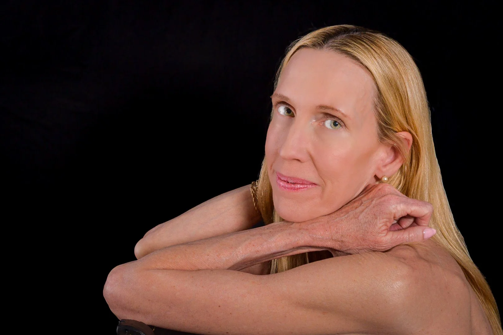 A woman with blonde hair and blue eyes resting her head on her folded arms, looking into the camera against a black background.