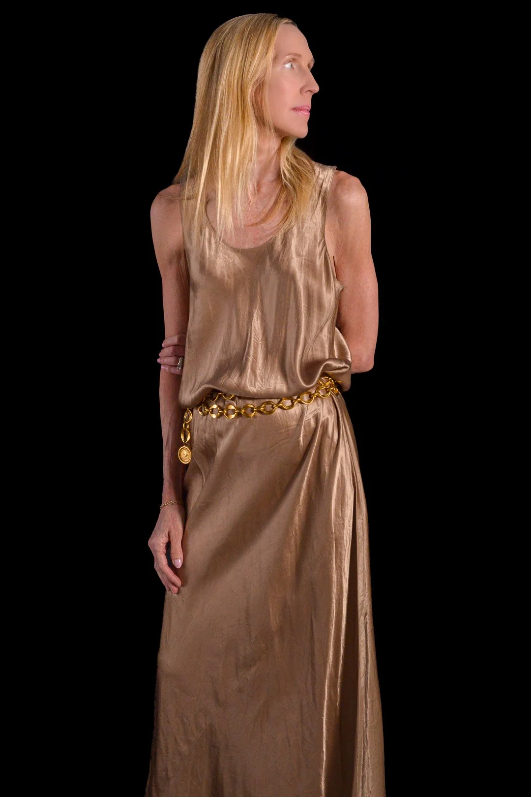 A woman with long blonde hair wearing a shiny gold silk sleeveless dress and a gold chain belt, standing against a black background.