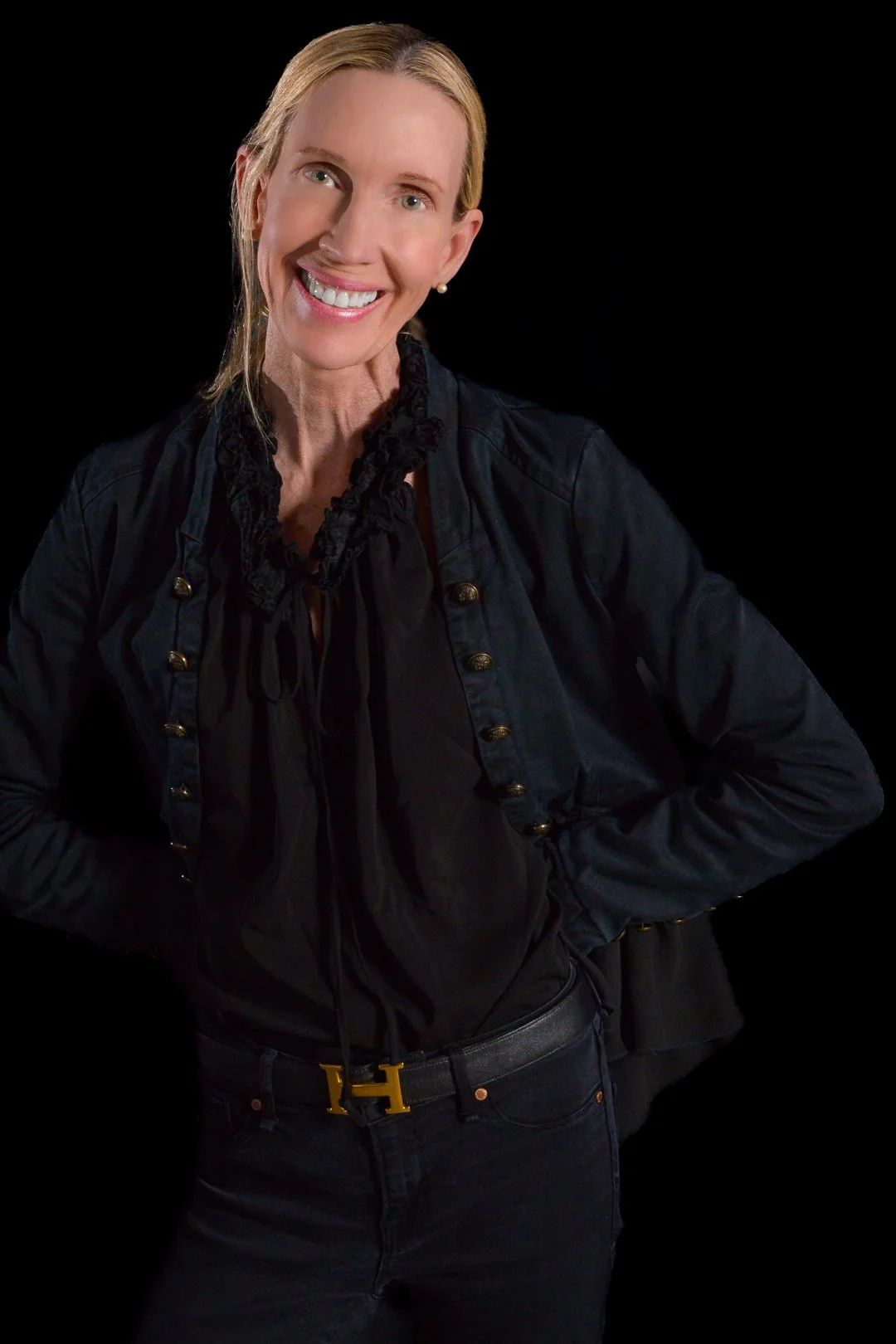 A smiling woman with blonde hair, wearing a black jacket with gold buttons and a black blouse with lace details, posing against a black background.