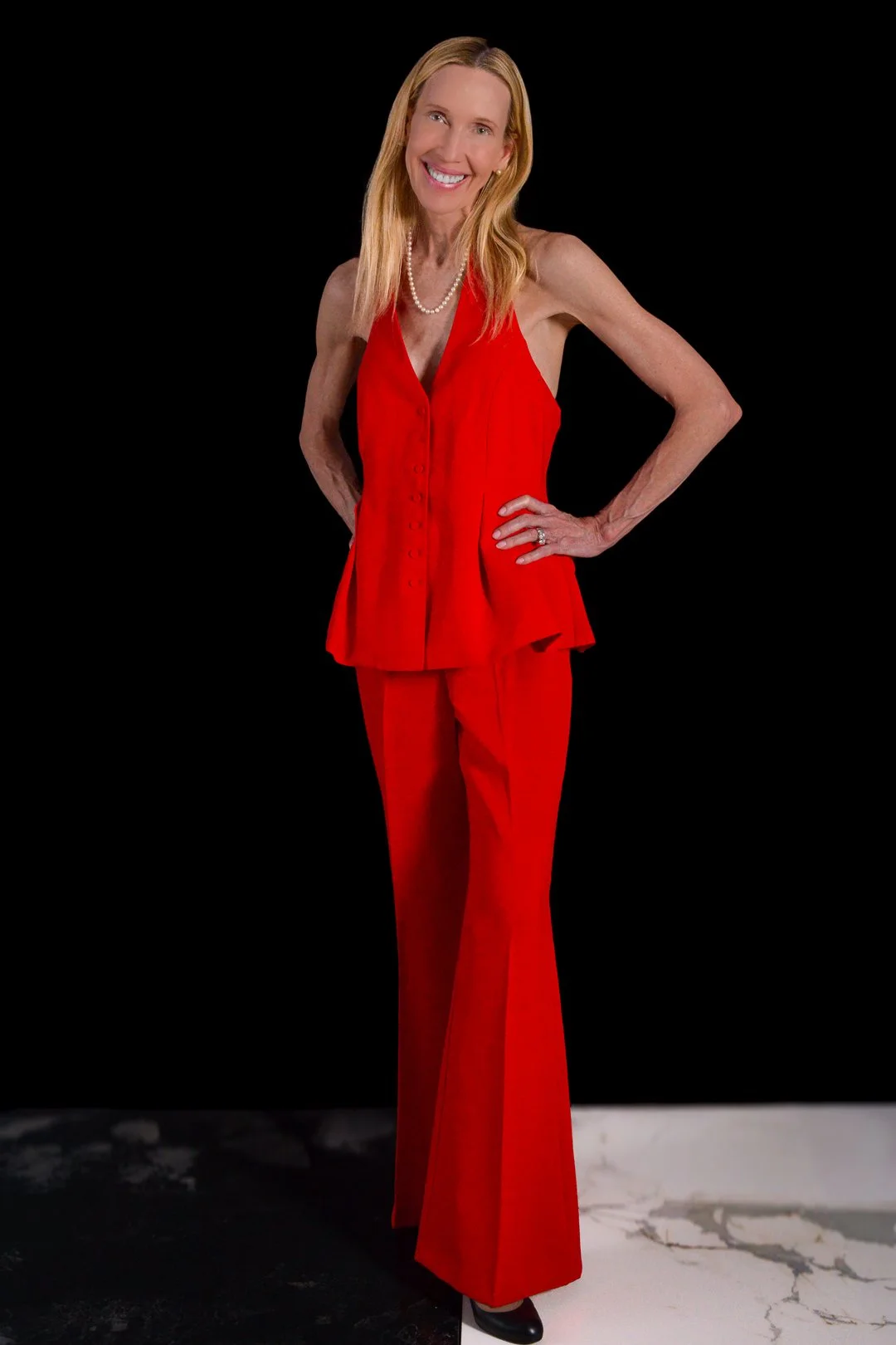 A woman with long blonde hair, wearing a red sleeveless top and matching wide-leg pants, smiling and posing with her hands on her hips against a black background.