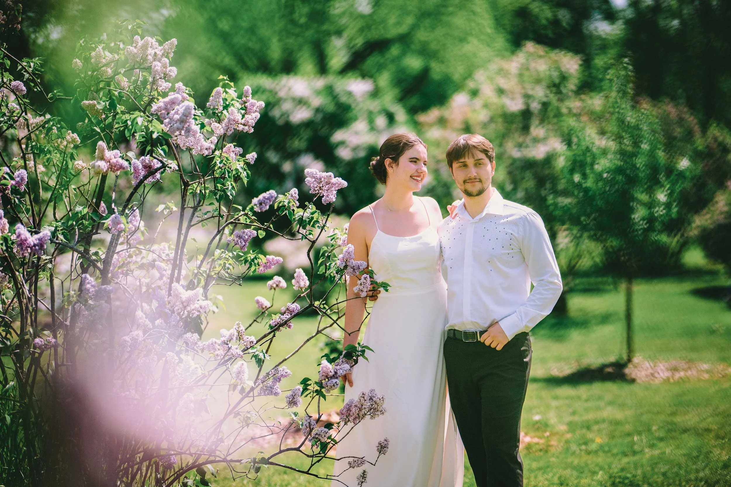 A romantic portrait of a couple standing amidst blooming lilac bushes, surrounded by vibrant greenery and soft light. This serene outdoor scene beautifully captures the joy and elegance of their love.
