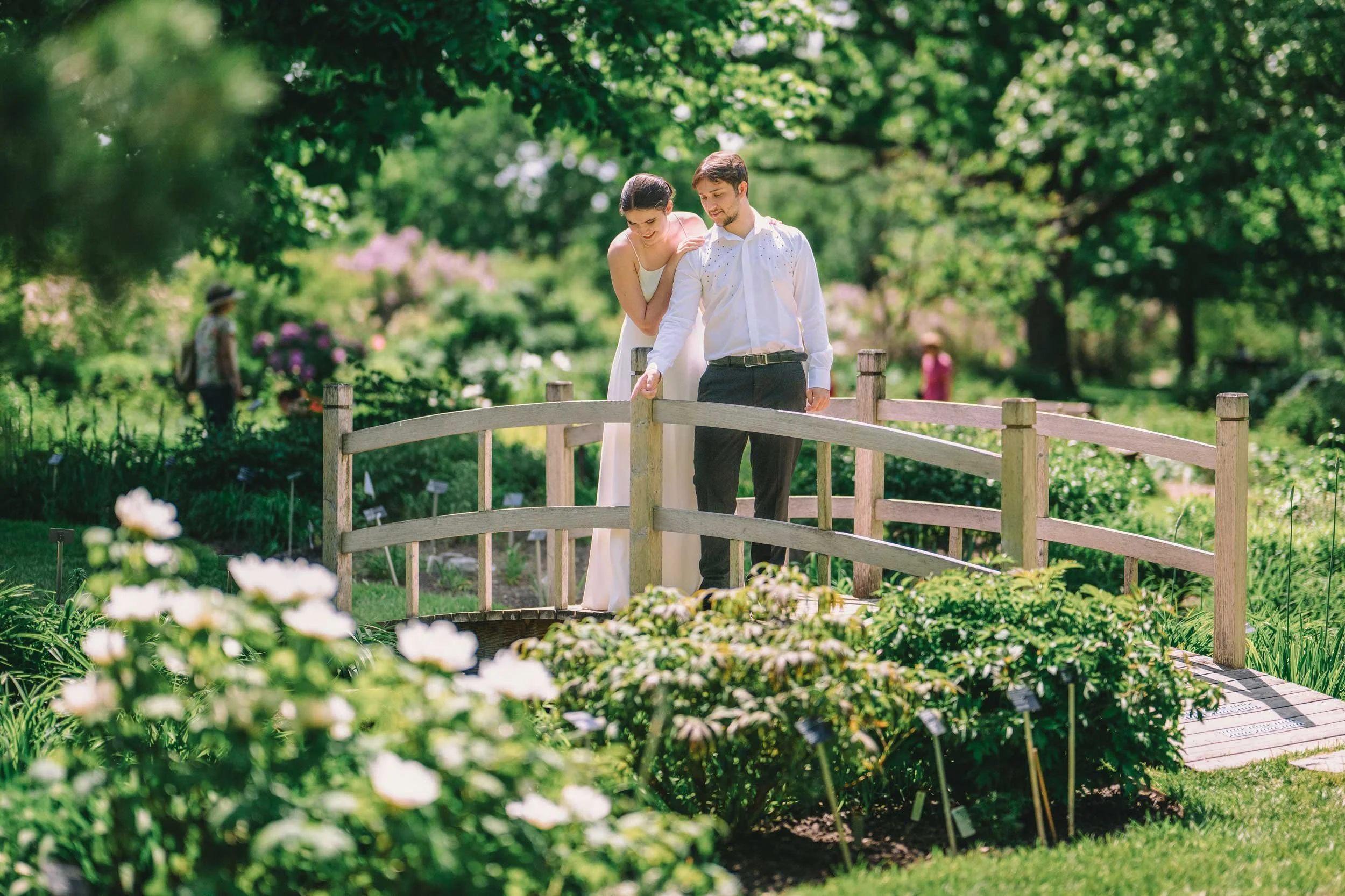 A romantic outdoor portrait of a couple standing on a rustic wooden bridge amidst a vibrant garden. Surrounded by blooming flowers and lush greenery, this serene moment captures the beauty of nature and love.