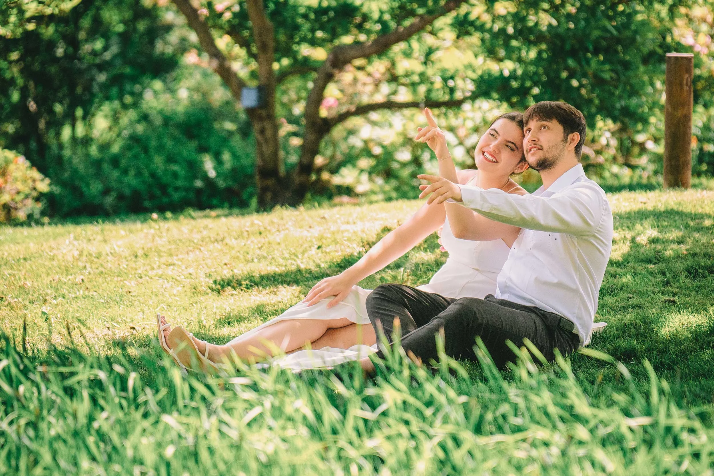 A cheerful outdoor engagement photo of a couple sitting on the grass under the shade of a tree. Their playful expressions and relaxed poses create a warm and romantic atmosphere, surrounded by lush greenery.