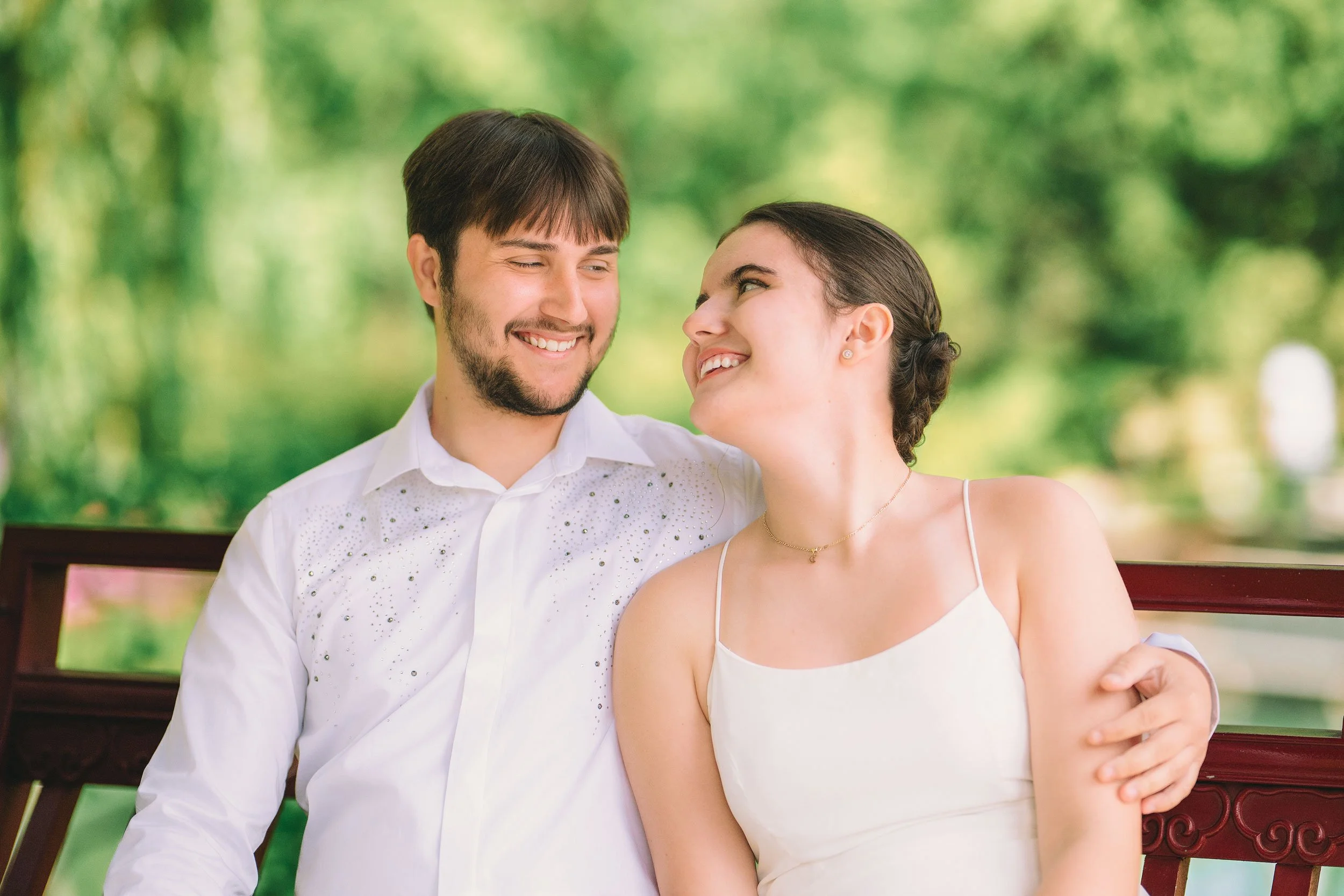 A joyful engagement portrait of a couple sharing a loving moment on a bench, surrounded by lush greenery. This natural light photo captures their happiness and connection in a serene outdoor setting.