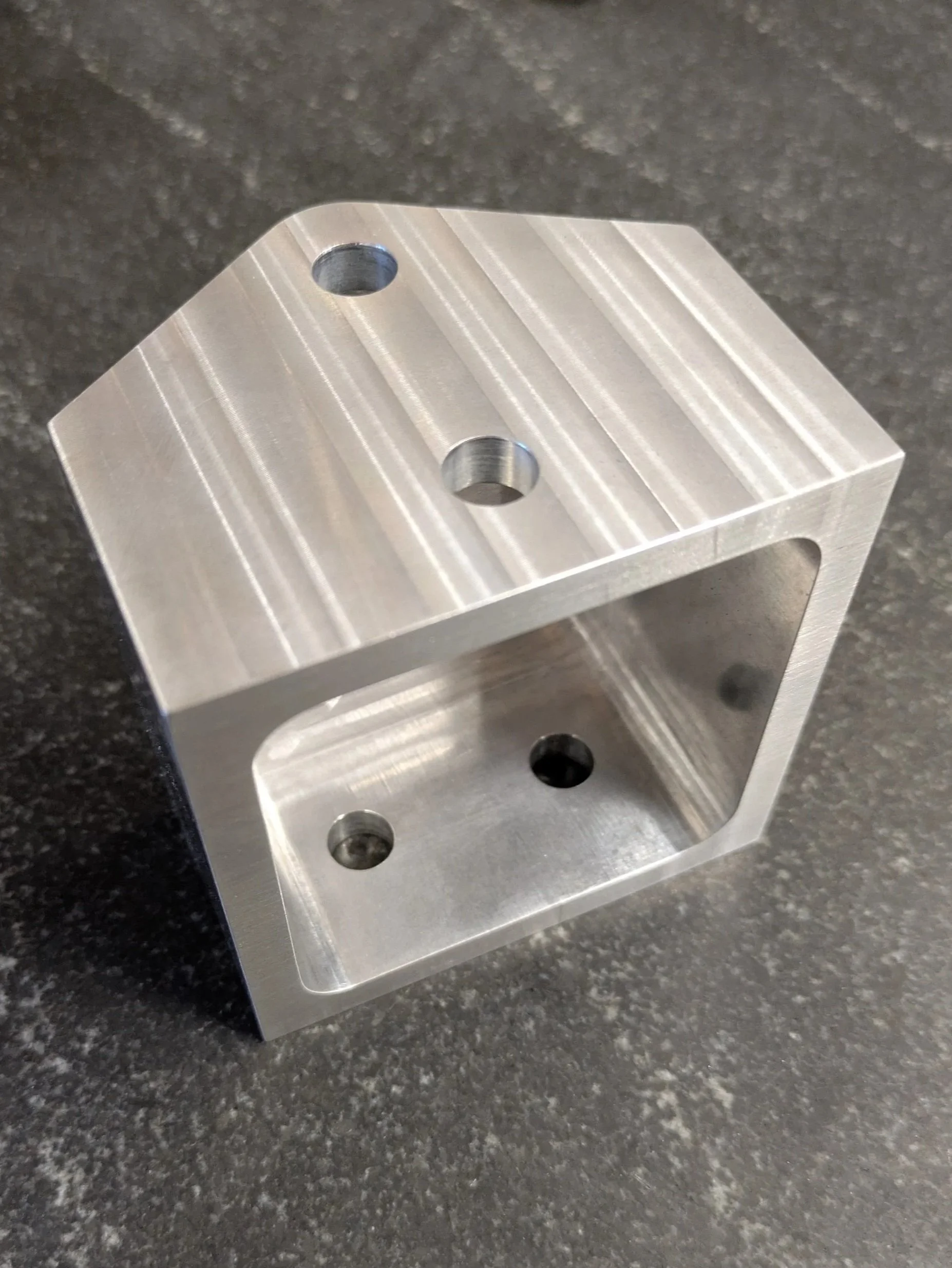 A close-up image of a silver aluminum block with threaded holes, placed on a black speckled countertop.