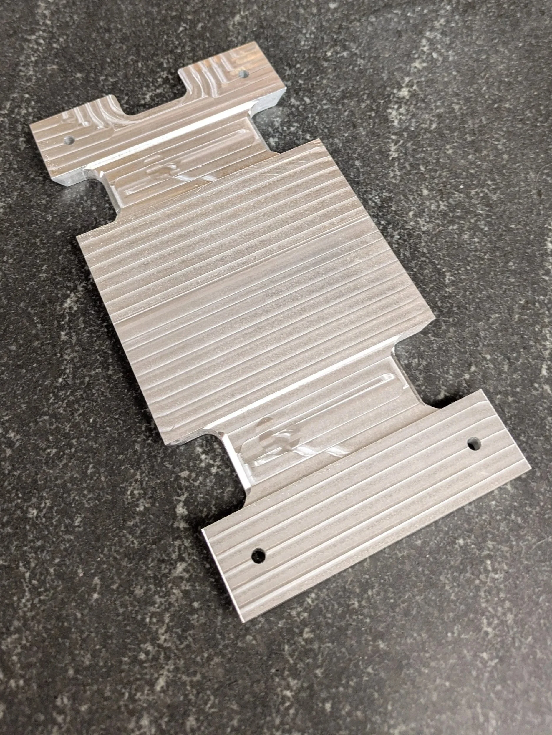 A metal car bumper reinforcement bracket with a ridged surface, placed on a dark speckled countertop.
