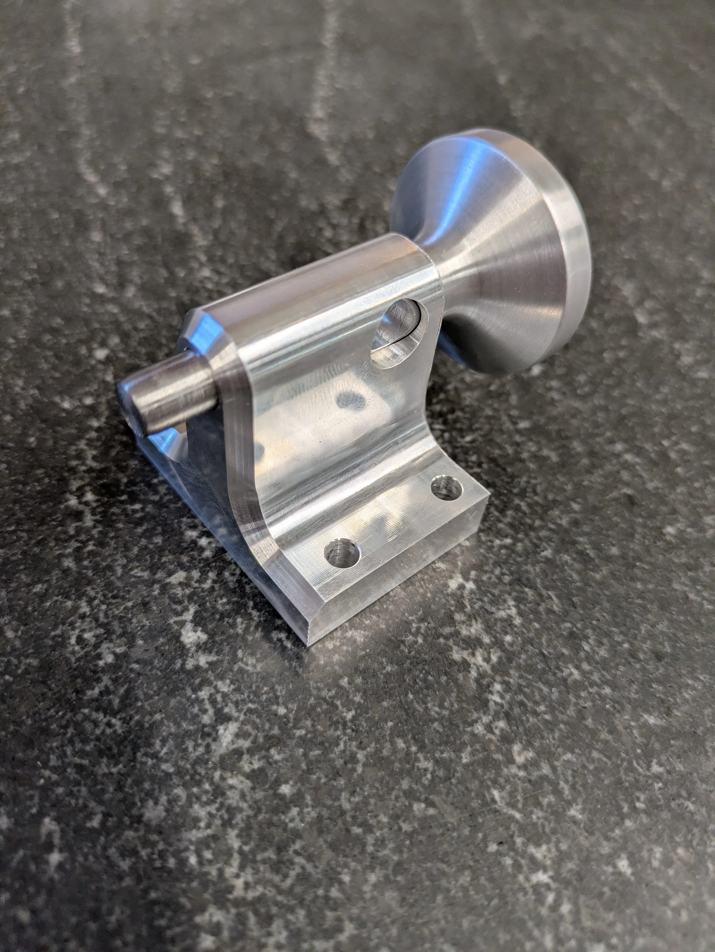 Close-up of a metallic, machined part with a cylindrical projection, holes, and a slotted hole, resting on a dark, speckled surface.