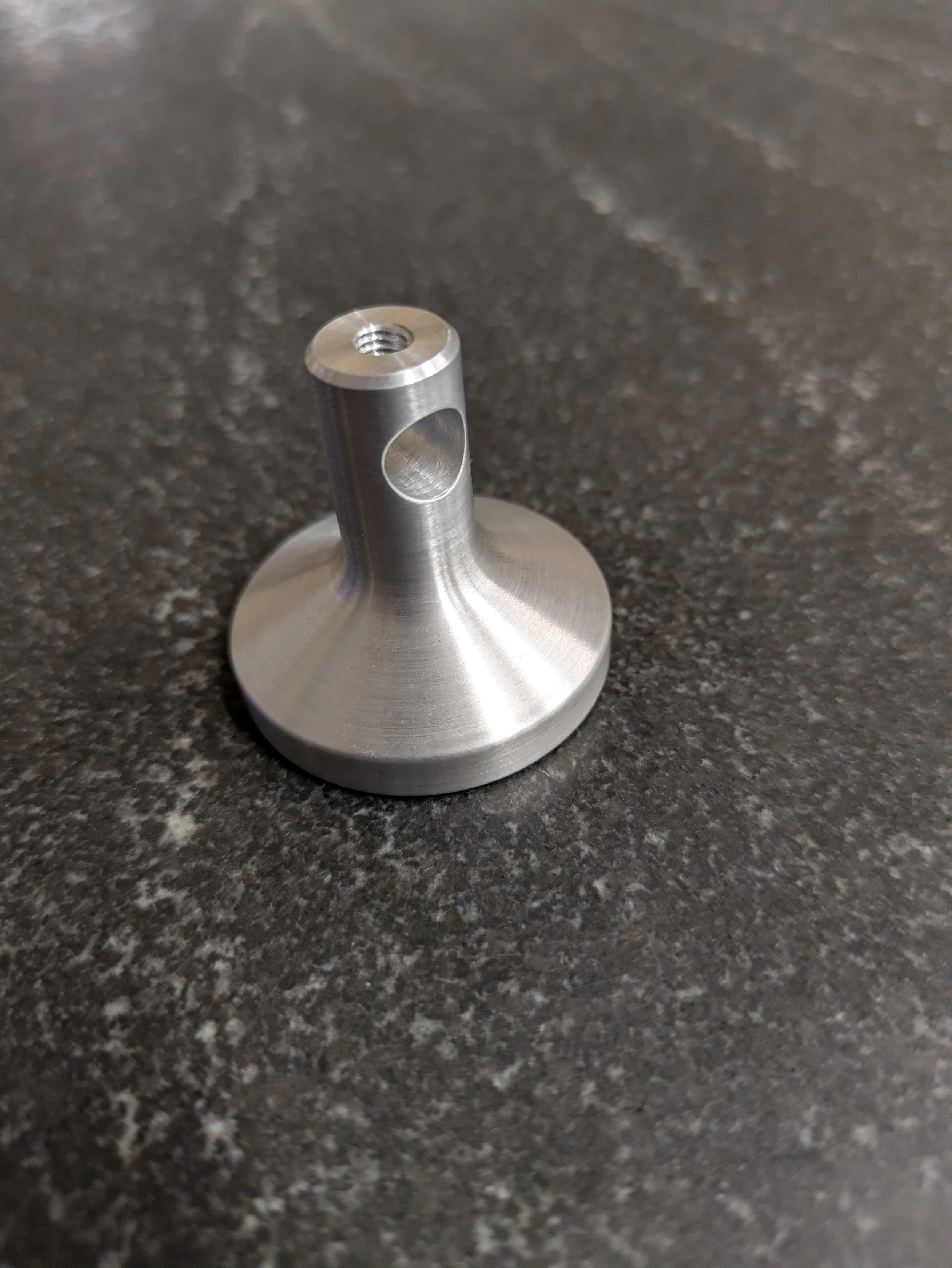 A small metallic mechanical component with a cylindrical shape, featuring a threaded hole on top and a circular opening on its side, positioned on a dark, speckled surface.