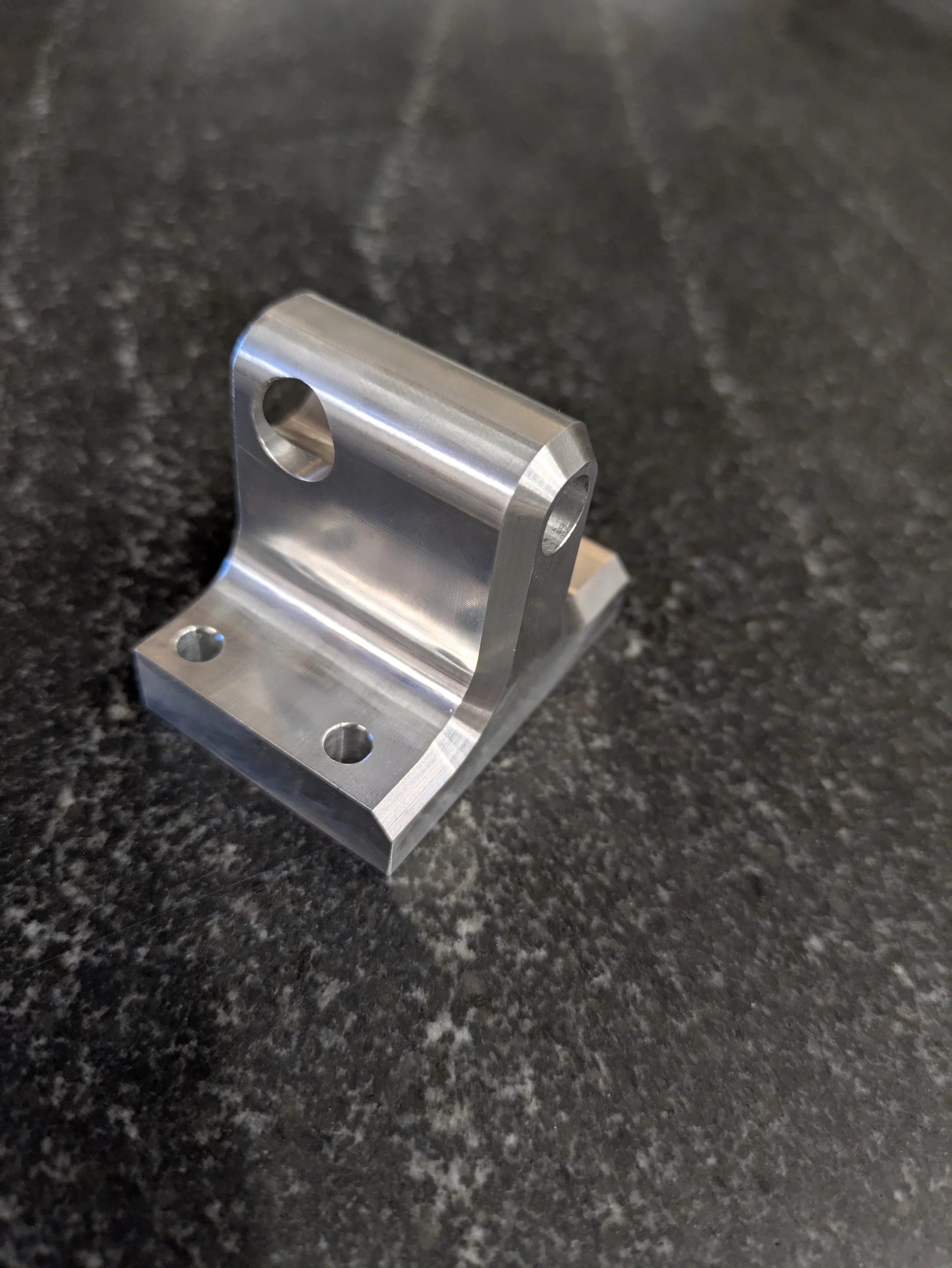 A small, silver, machined metal bracket with multiple holes resting on a dark granite countertop.