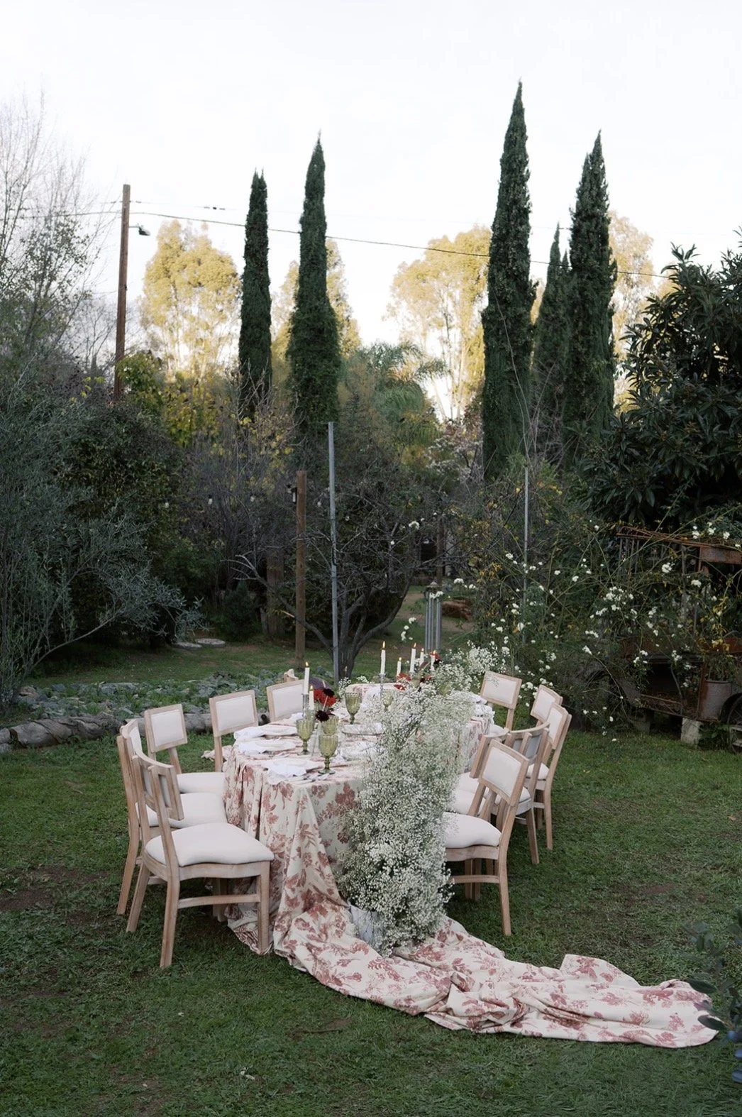 A round dining table decorated with a floral tablecloth, white chairs, candles, and flowers, set outdoors in a garden with lush greenery and tall trees during daylight.