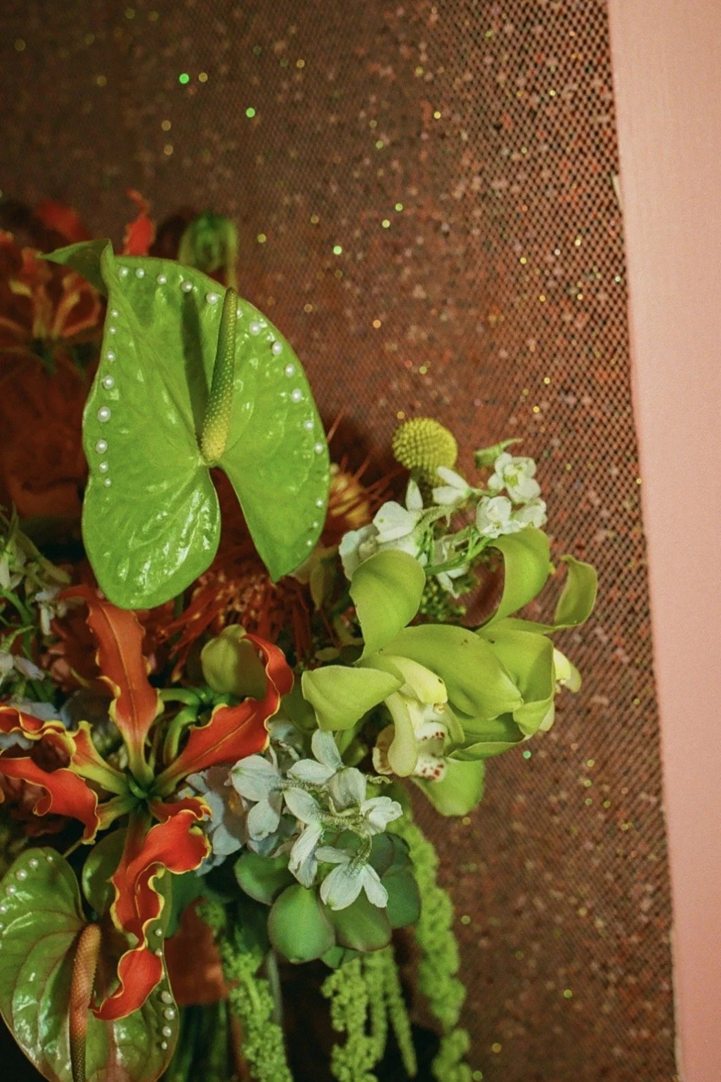 Artificial flower arrangement with green, red, and white flowers and leaves, against a glittery rose gold background.
