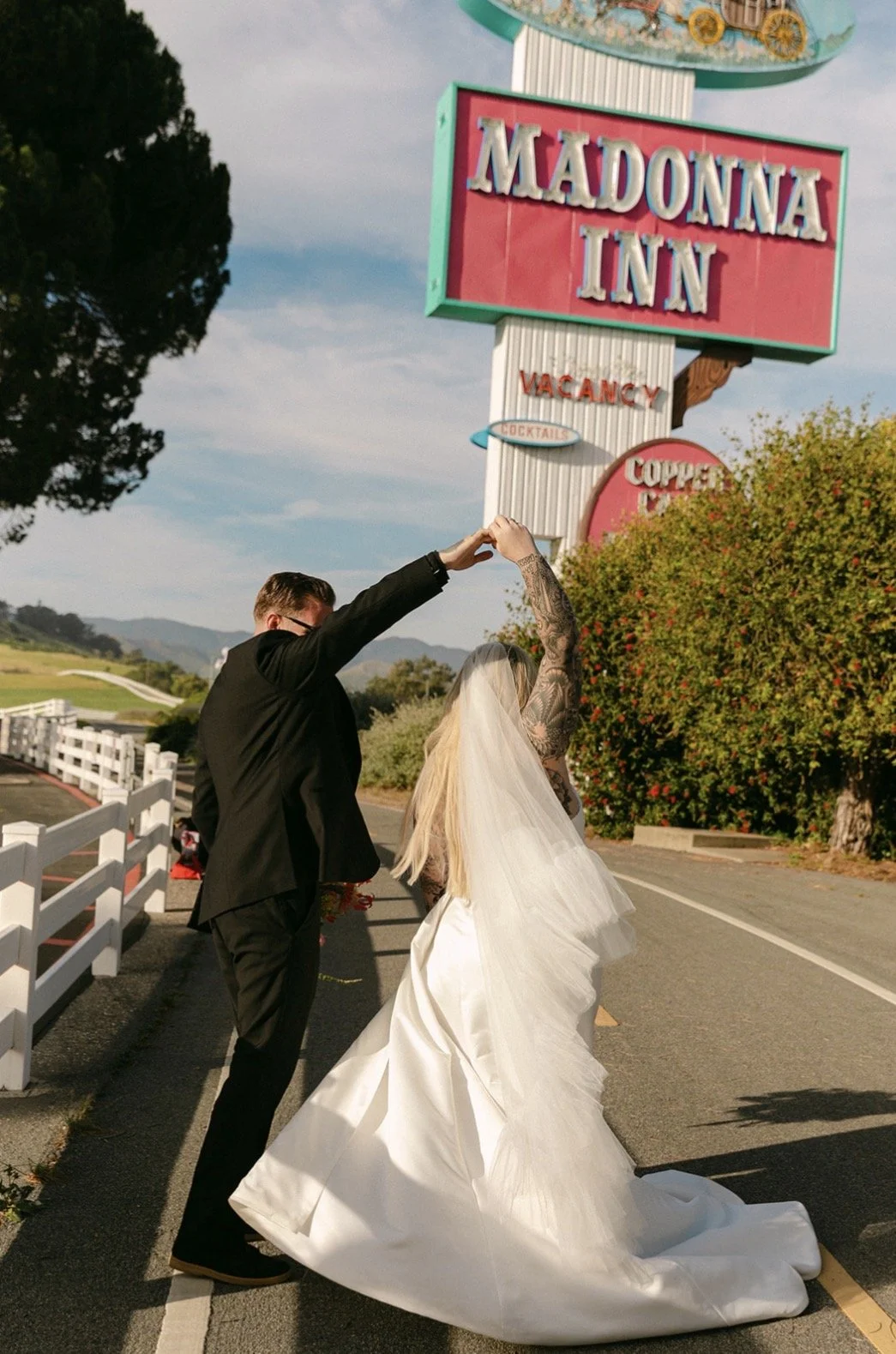 A couple in wedding attire dancing on the road outside the Madonna Inn sign, with a white fence and trees in the background.