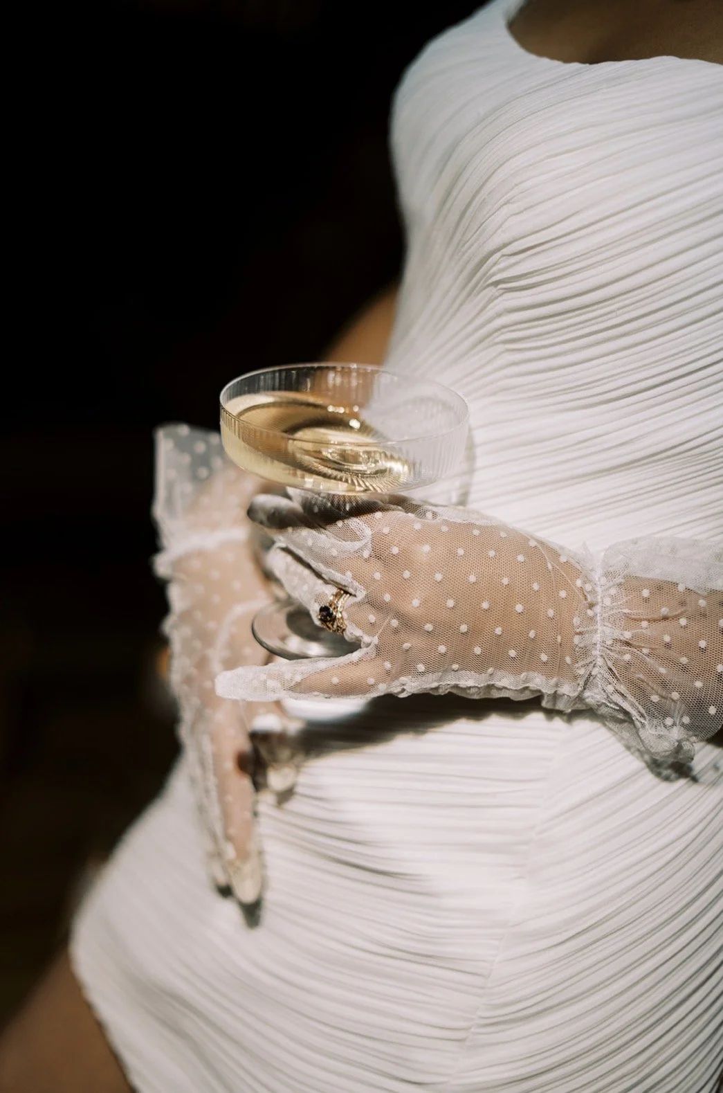 Person wearing white dress and sheer polka dot gloves holding a glass of champagne.