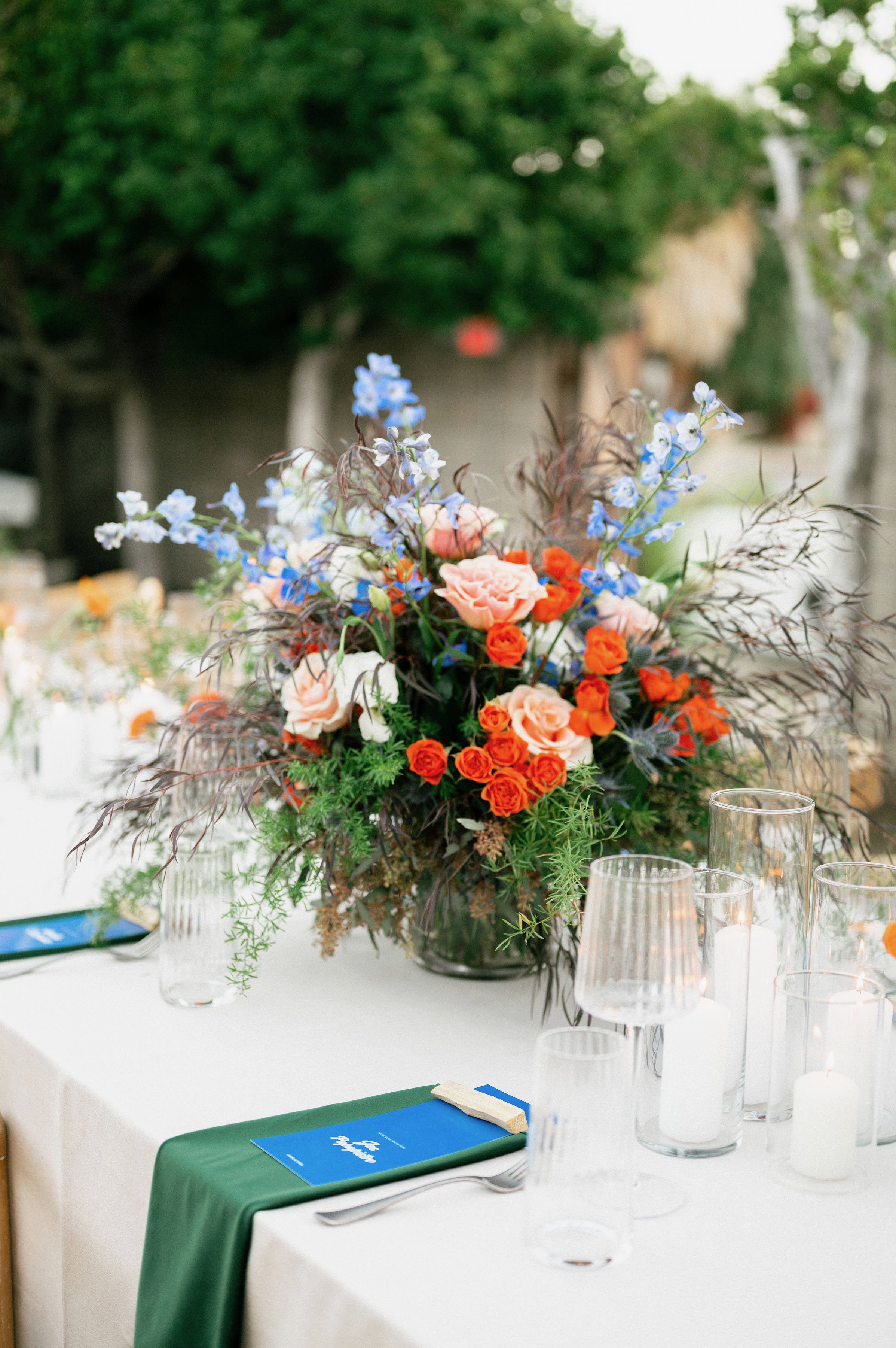 A large floral centerpiece with pink roses, orange spray roses, and blue flowers on a dining table decorated with a green table runner, glassware, and candles, set outdoors.